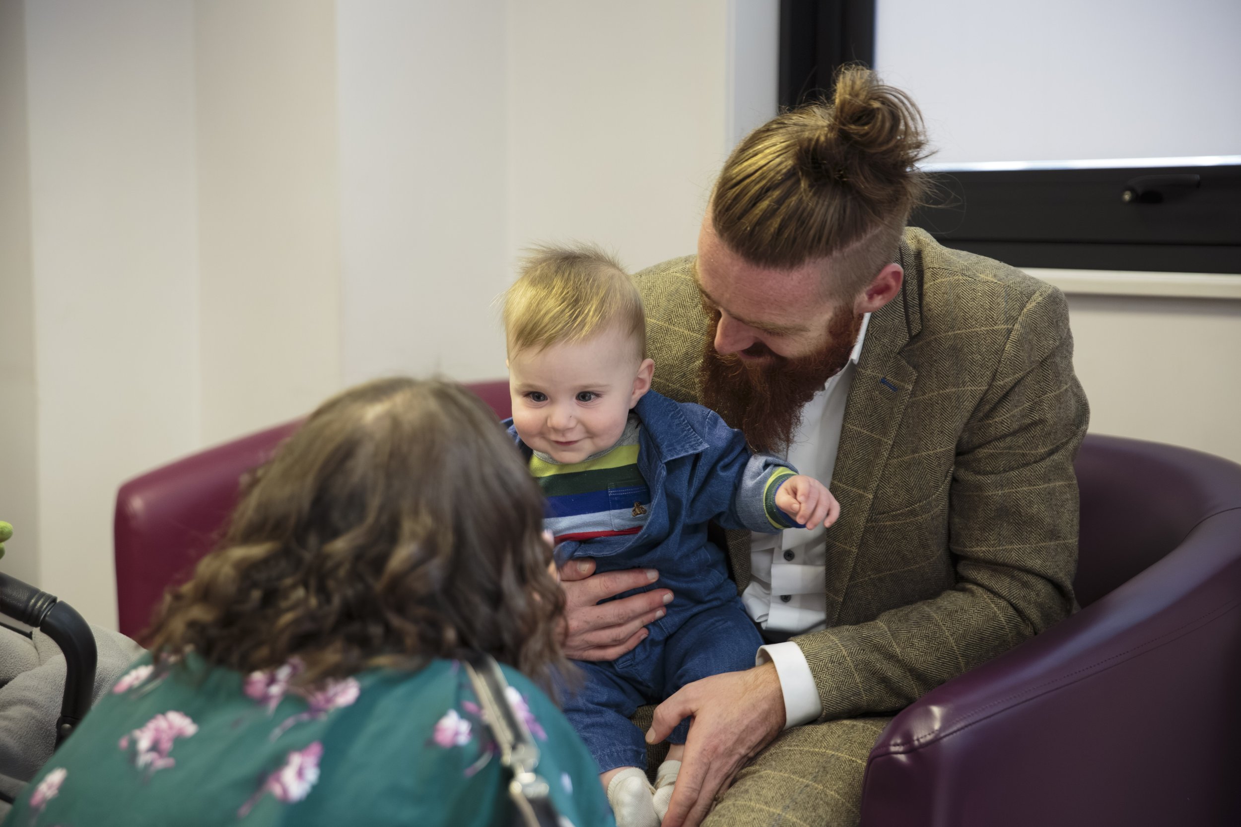 A man with a red beard wearing a brown checkered blazer is sitting in a waiting room holding a young boy with blonde hair, wearing a blue jacket, on his lap. Two children are in the foreground, one with brown hair and part of another with light-color
