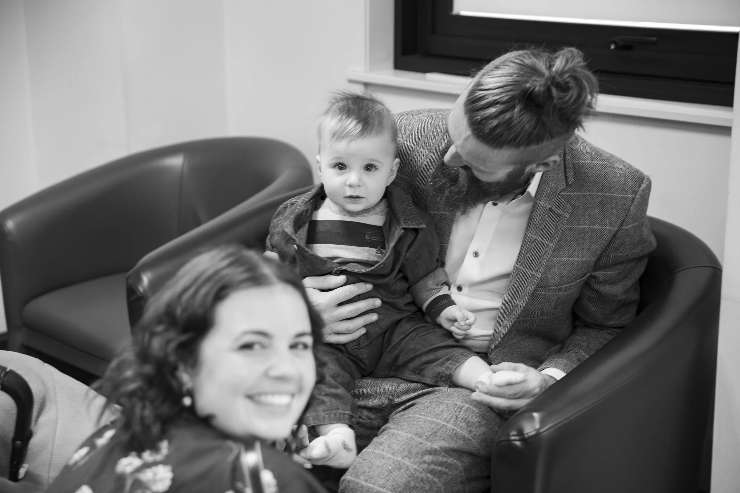 A man with in glasses and a beard sitting in a room with two children, one in his lap and the other in a stroller, during daytime.