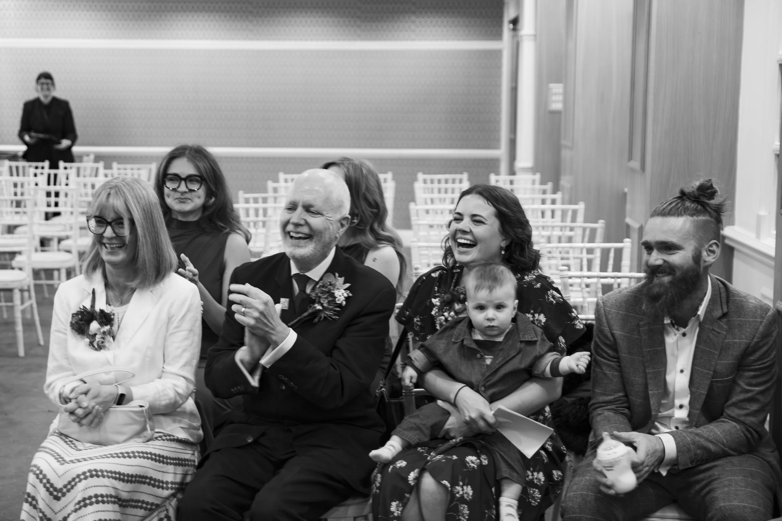 A black and white photo of a group of people sitting together, laughing, at an indoor event. There are chairs in the background, and a woman stands in the back.