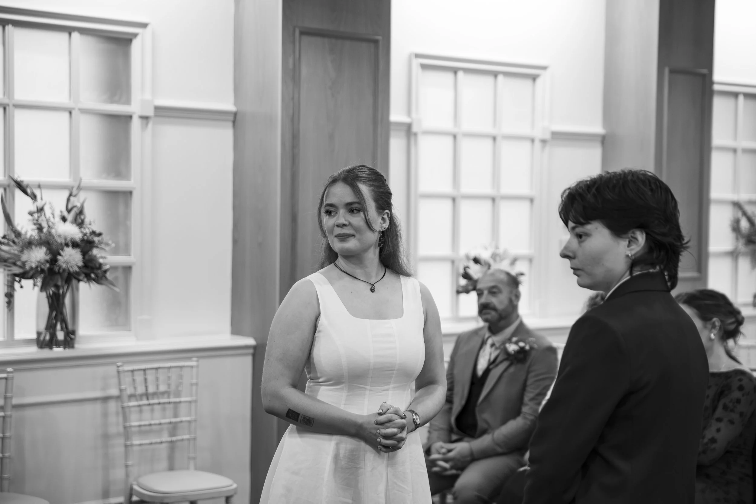 A woman in a white dress stands in a room with wood-paneled walls and large windows. She appears to be at a formal event, possibly a wedding, and is looking to the side with a slight smile. There are other people seated around her, including a man in