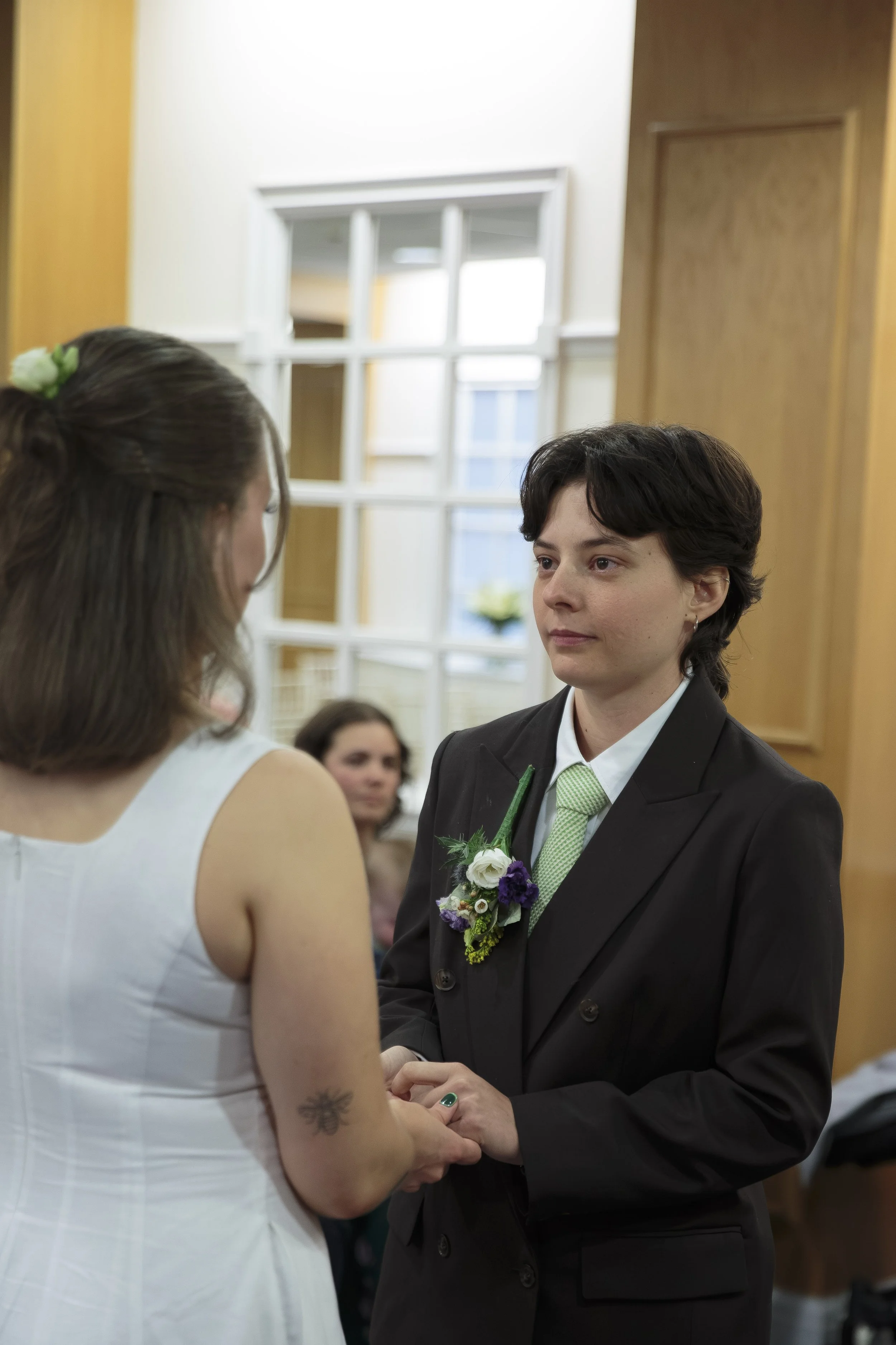 A close-up of an engaged couple exchanging wedding vows indoors. The woman wears a white dress with a tattoo on her arm, and the man is in a dark suit with a green tie and a boutonniere of flowers. They hold hands and look into each other's eyes as p