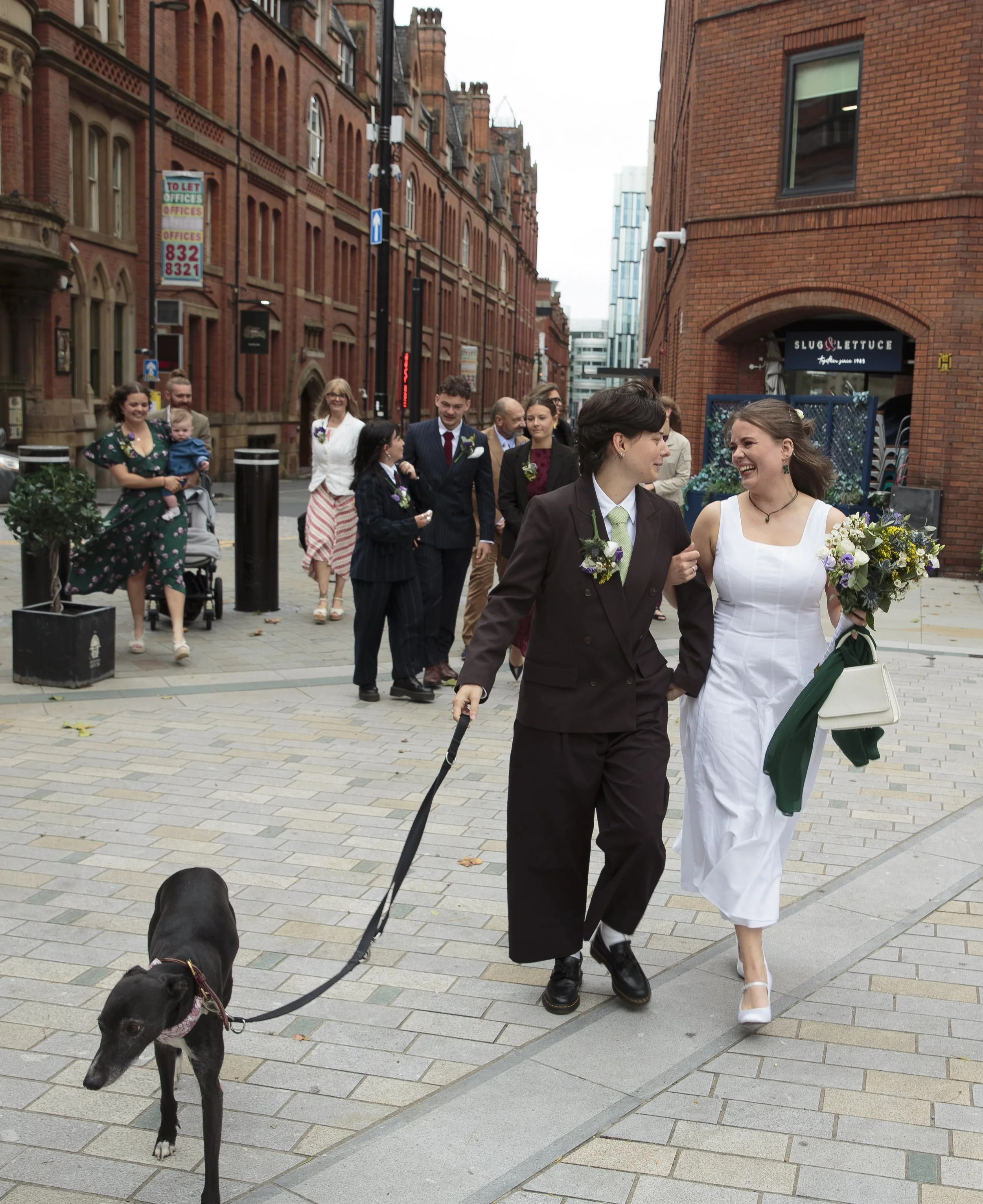 group of people walking along a city street, including a woman in a white dress holding a bouquet and a woman in a brown suit walking a black dog on a leash.