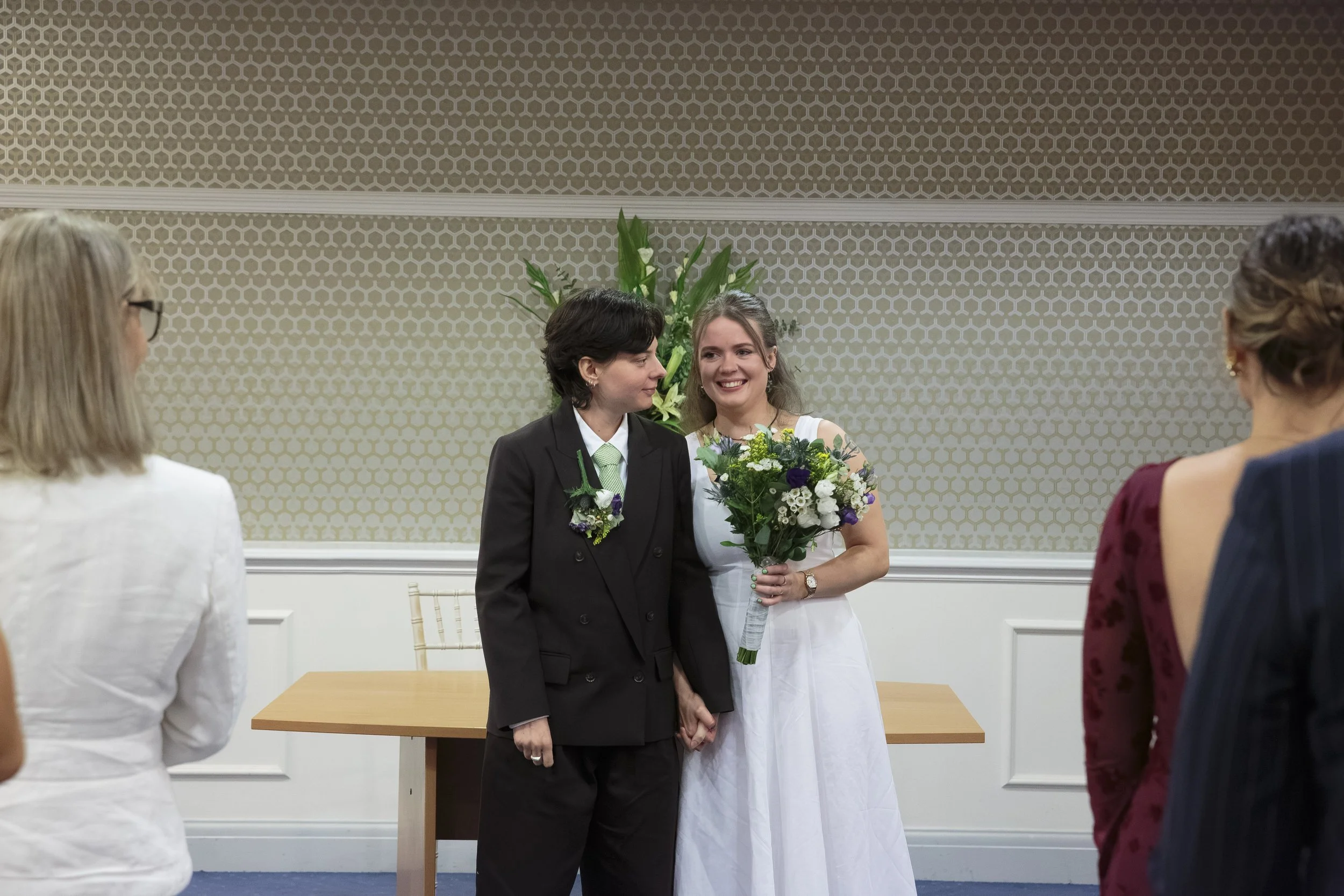 Two women stand together during a wedding ceremony, holding hands, with a bouquet of flowers. One woman is wearing a white dress and the other is in a dark suit. They are smiling and facing each other, surrounded by three women watching.