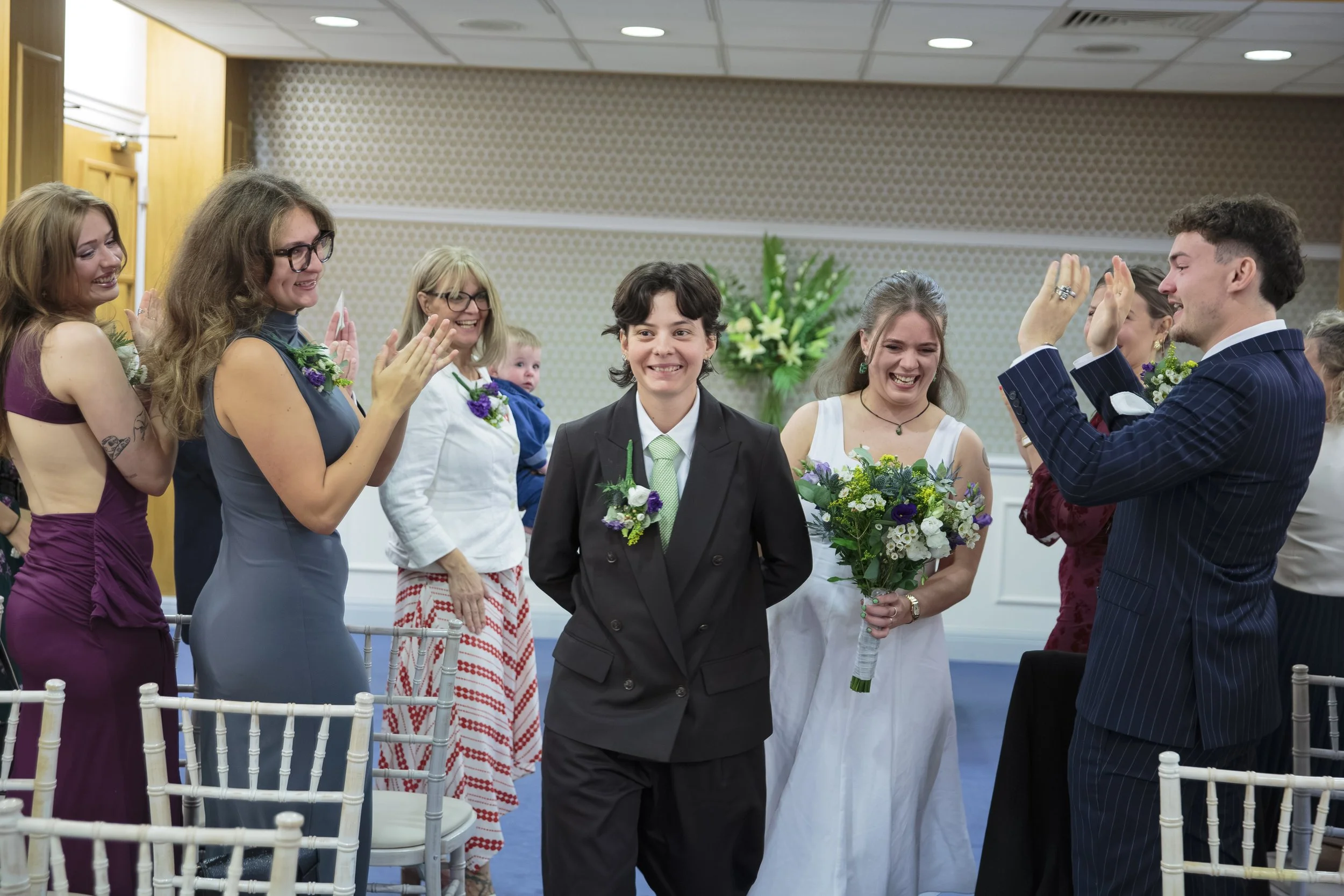 Group of people at a wedding, with a woman in a purple dress, a woman in a blue dress, a woman in a white dress, and a man in a striped suit, all smiling and giving high-fives to a joyful young man in a black suit holding a bouquet, in a decorated in