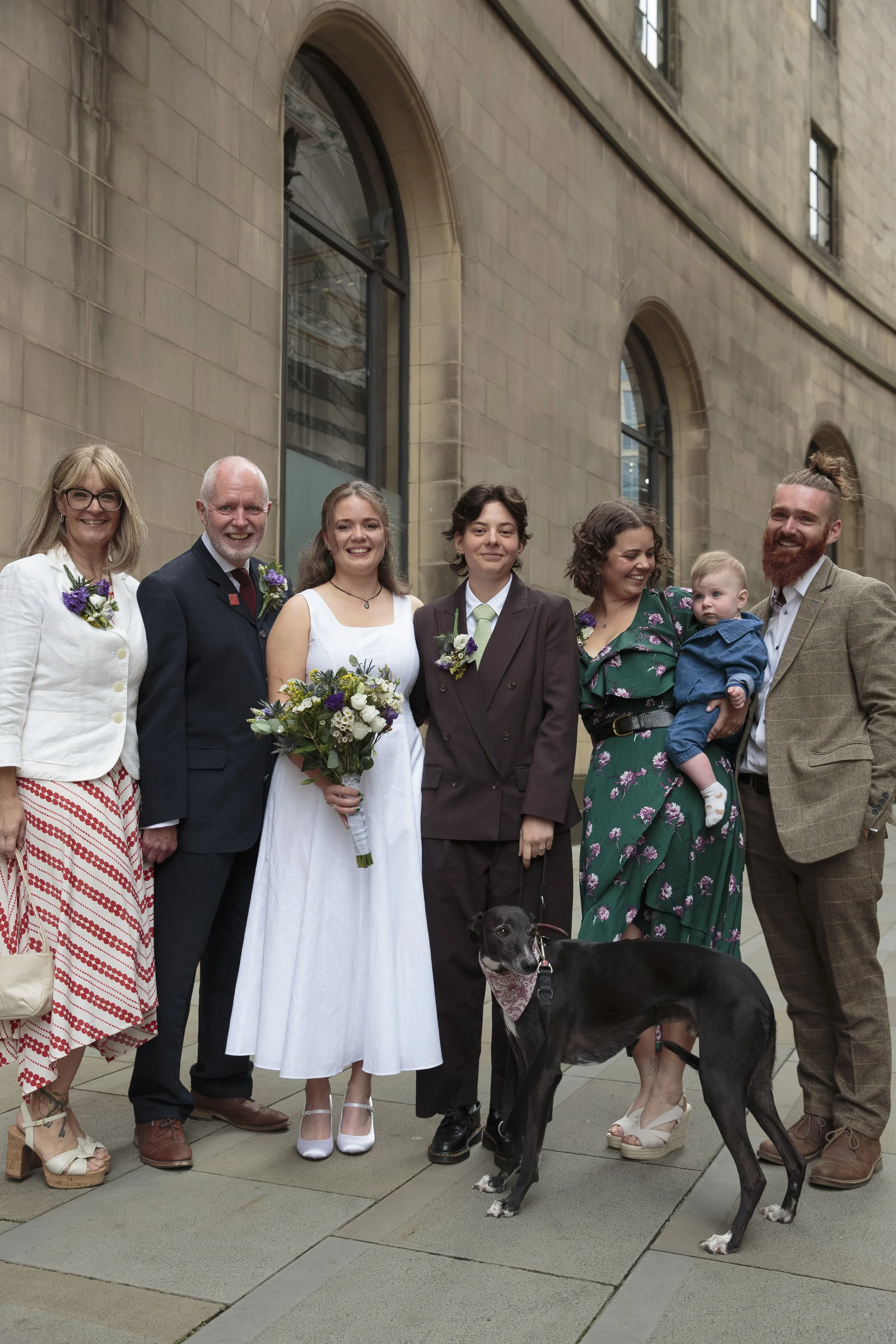 Group of seven people and a dog standing on a city sidewalk in front of a stone building. The group includes men and women, some holding flowers, and a young child being held by a woman. Everyone is dressed in semi-formal attire, with some wearing su