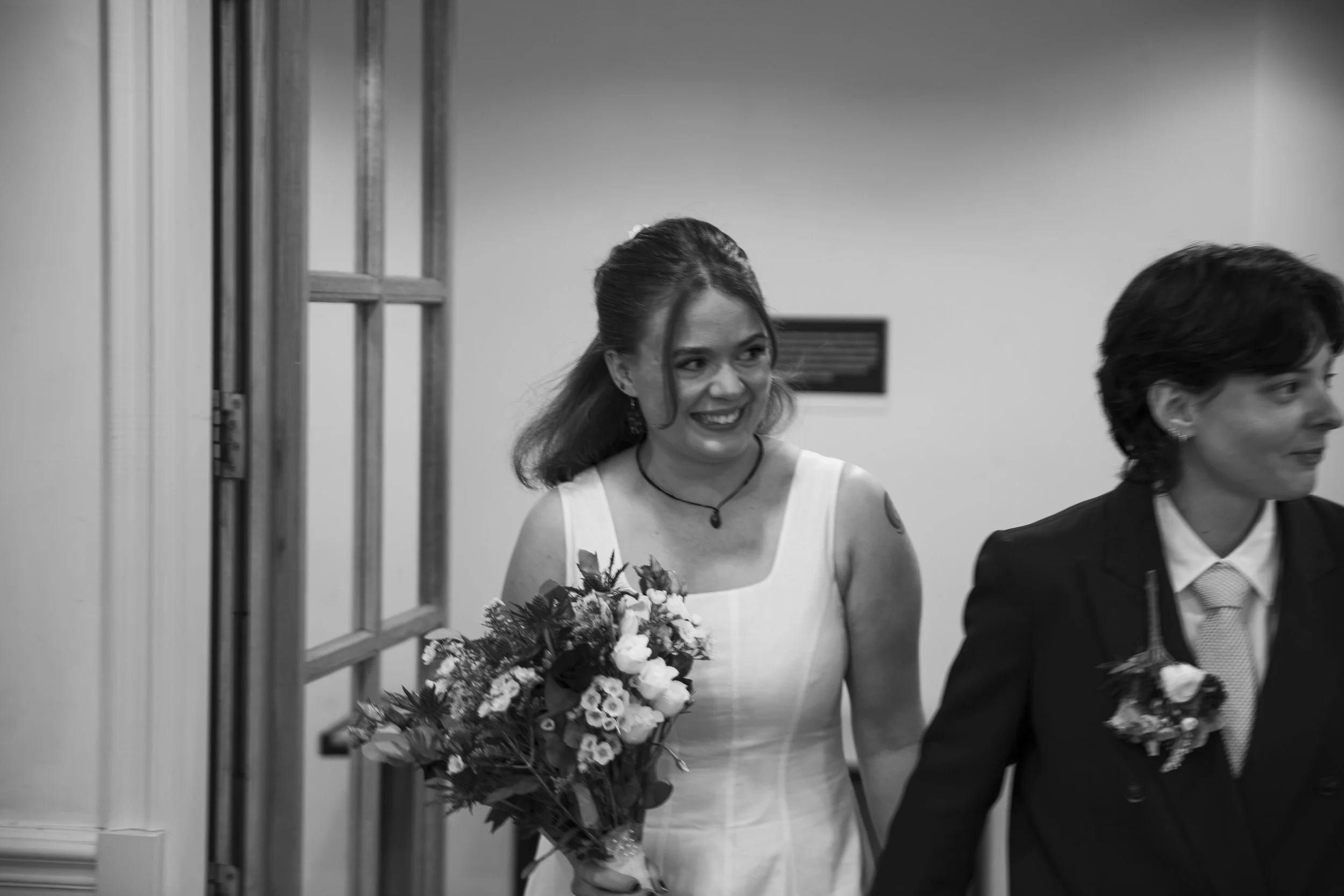 A woman in a white dress holding a bouquet of flowers, standing next to a woman in a dark suit, both smiling at an event or celebration.