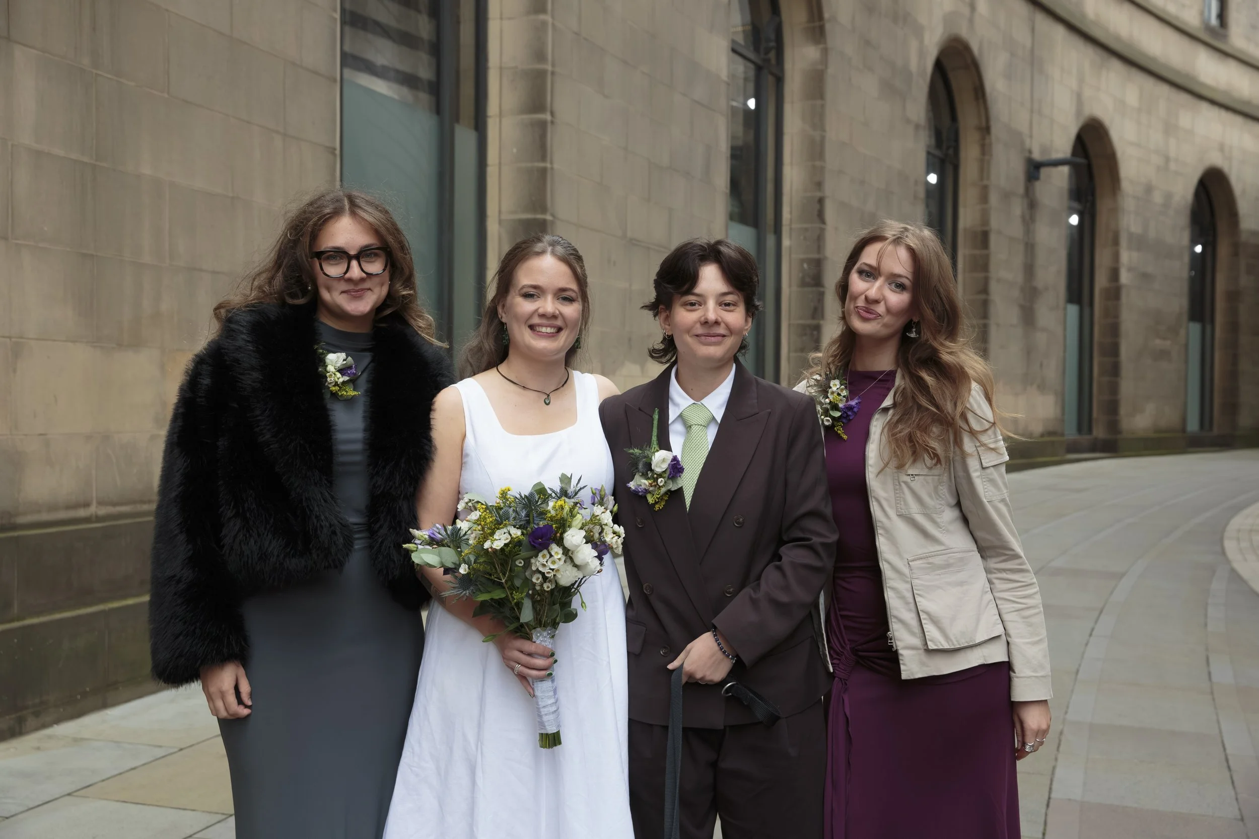 Four women standing together outside a stone building, one woman in a white dress holding a bouquet of flowers, others dressed in formal attire, smiling for the photo.