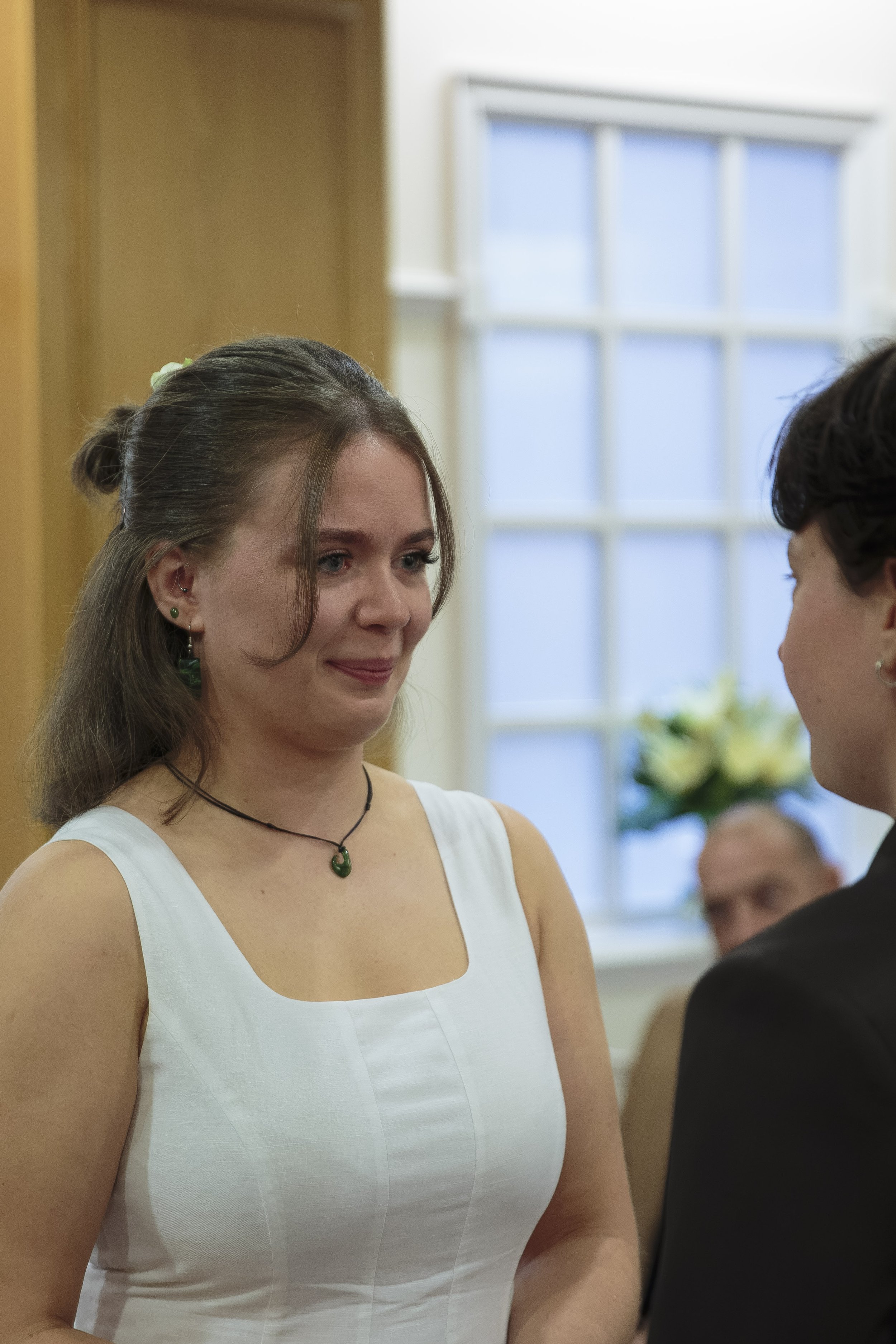A young woman with brown hair in a bun, wearing a white dress and green jewelry, looking at a young man in formal attire, during a wedding or similar ceremony in a bright room with a large window and a flower arrangement in the background.