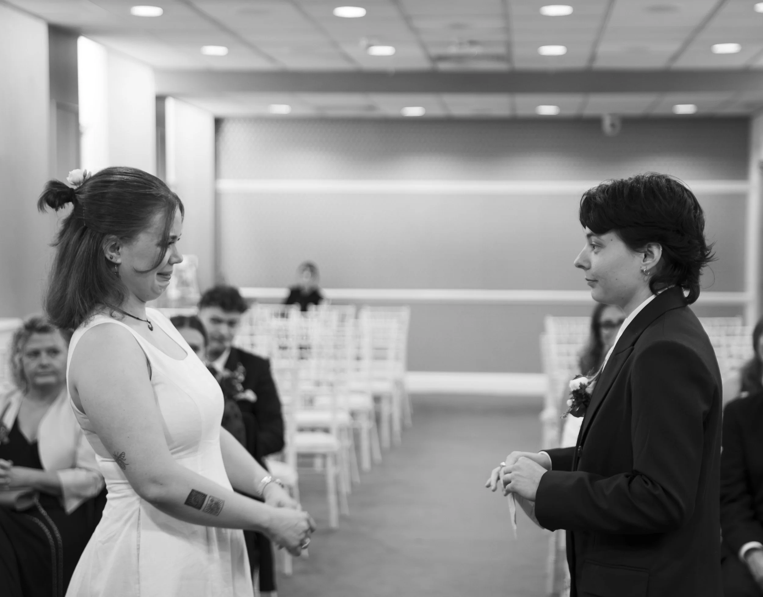 Two women standing face-to-face in a formal setting, holding hands, with several blurred people in the background, indoors with ceiling lights.