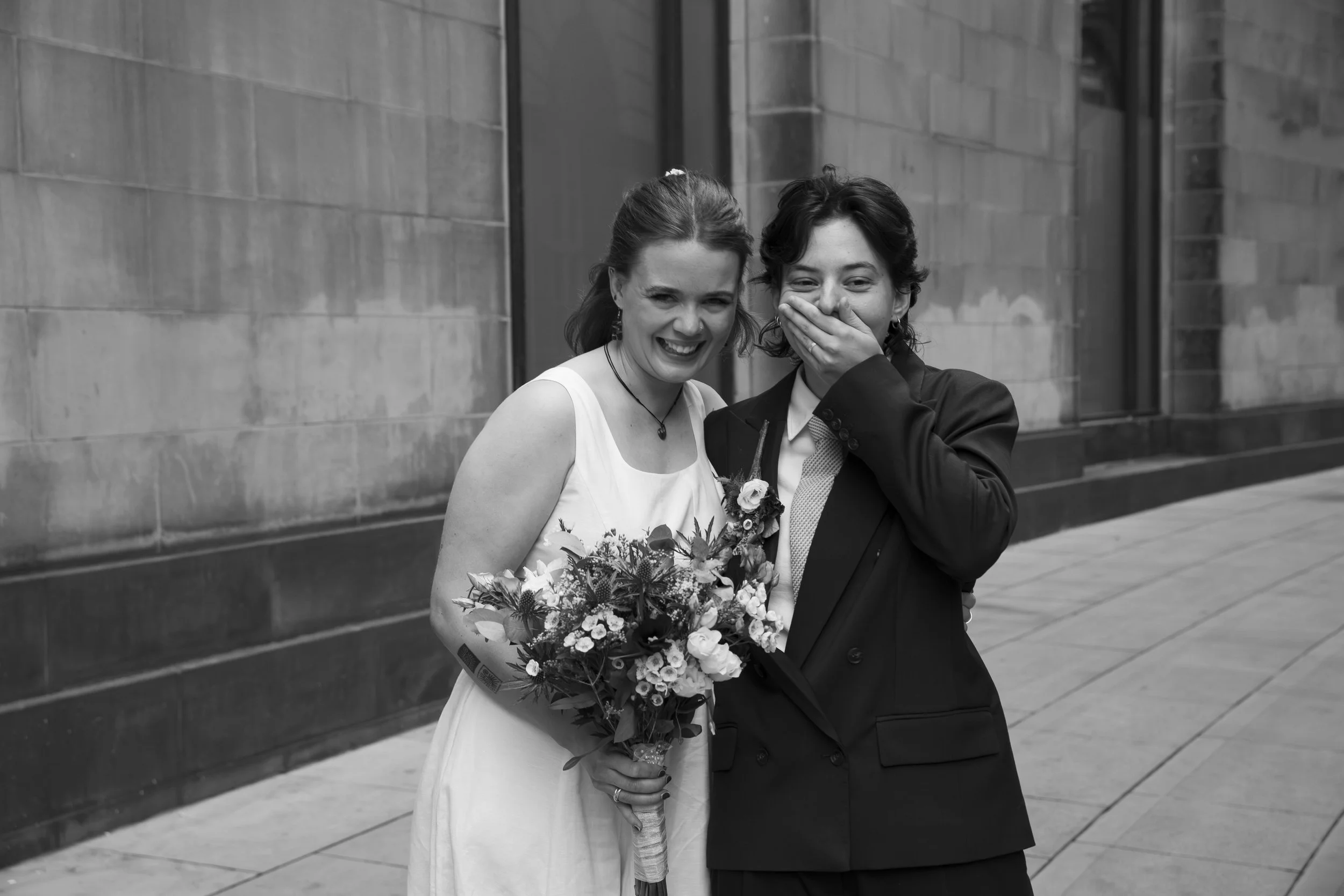 Two women, one wearing a wedding dress and holding a bouquet of flowers, and the other in a suit, are standing close together outdoors in an urban setting, smiling and covering her mouth with her hand.