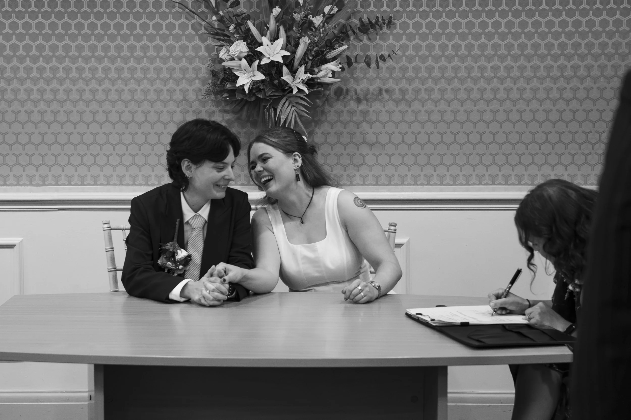 Two women sitting at a table, holding hands and smiling, during what appears to be a wedding ceremony. One woman is dressed in a black suit with a boutonniere, and the other in a white dress with a tattoo on her right shoulder and wearing earrings. A