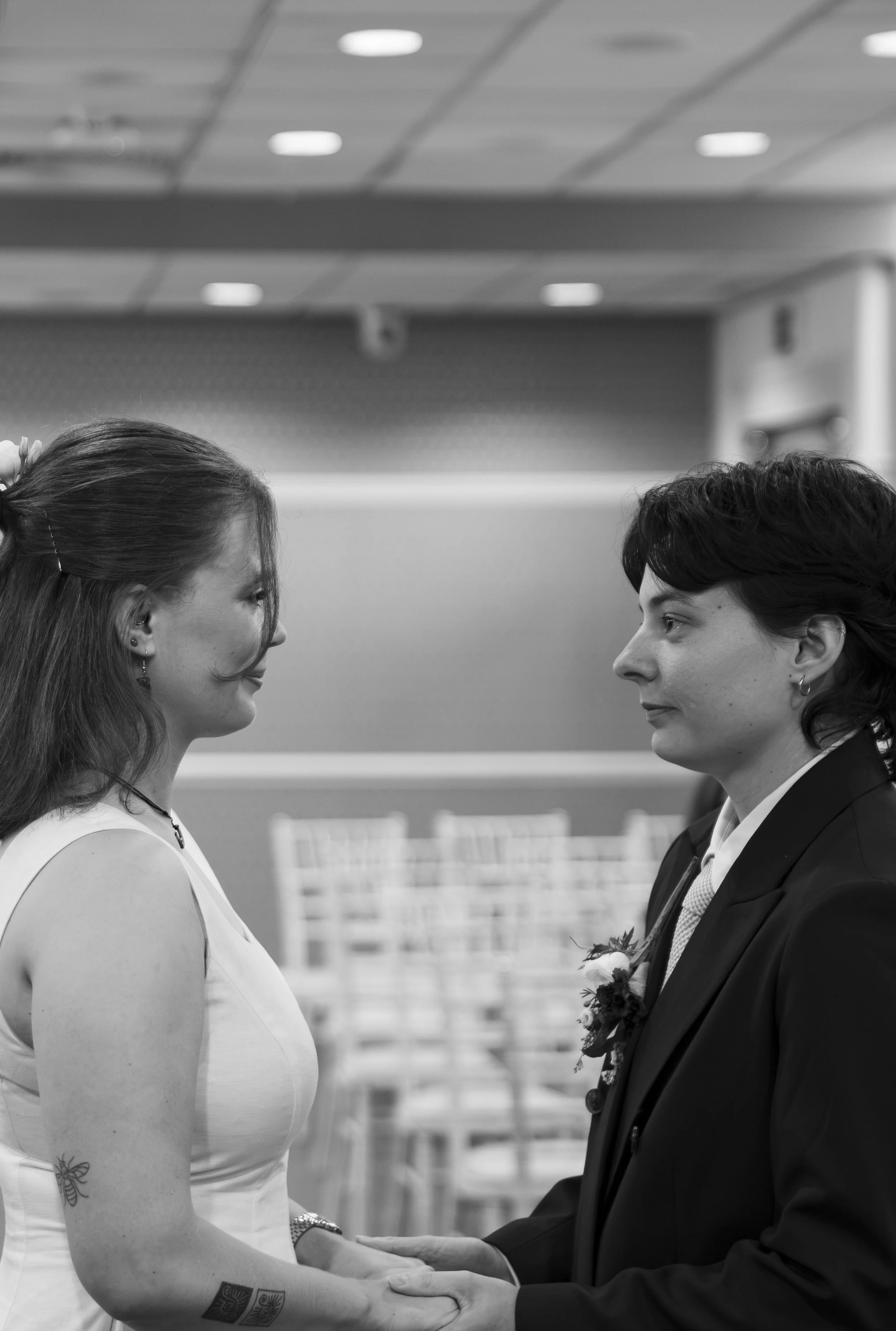 A woman and a man are facing each other and holding hands in a formal indoor setting, possibly a wedding or ceremony. The woman has long hair, earrings, and visible tattoos on her arm. The man has short hair, earrings, and is wearing a suit with a bo
