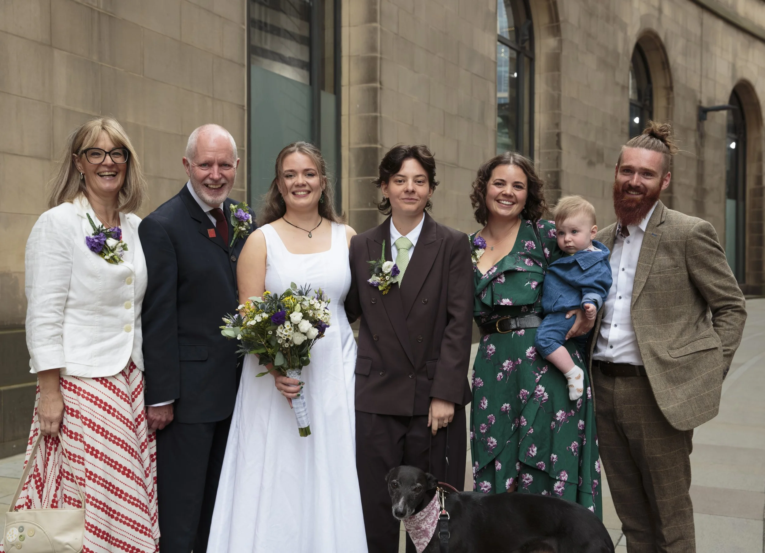 Group of seven people, four women, two men, and a child, standing outdoors in front of a stone building, smiling and dressed in formal and semi-formal clothing, with some wearing boutonnières and holding flowers, accompanied by a black dog.