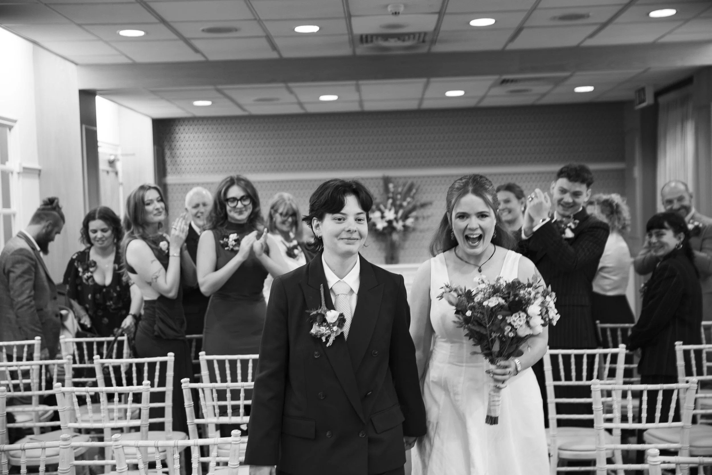 A group of people dressed formally at a celebration, including a woman in a wedding dress holding a bouquet and a man in a suit. They are smiling and clapping in a decorated indoor venue.