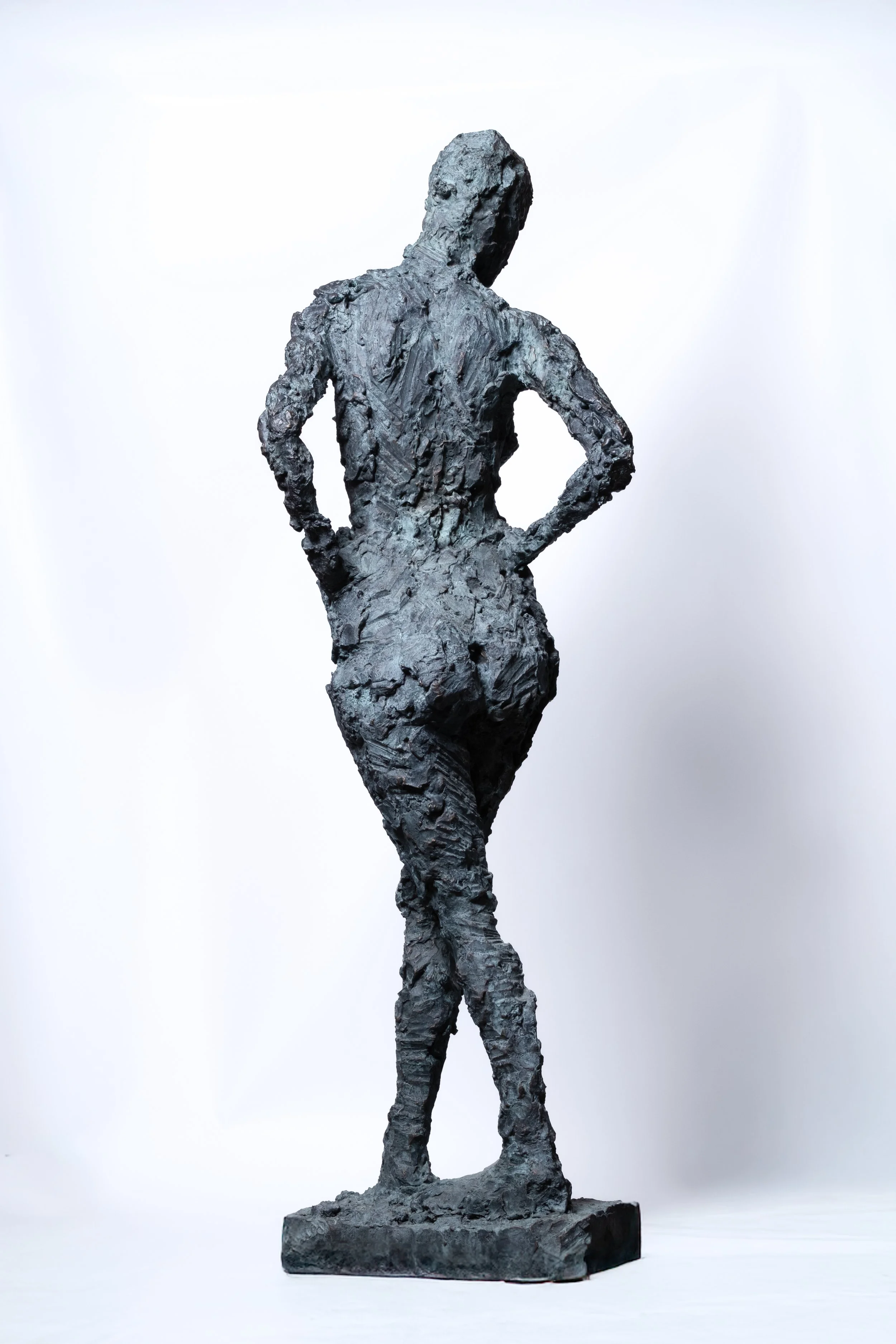 Eine dunkle, strukturierte Skulptur einer Frau, die mit den Händen in den Hüften steht und dem Betrachter den Rücken zuwendet, vor einem schlichten weißen Hintergrund.