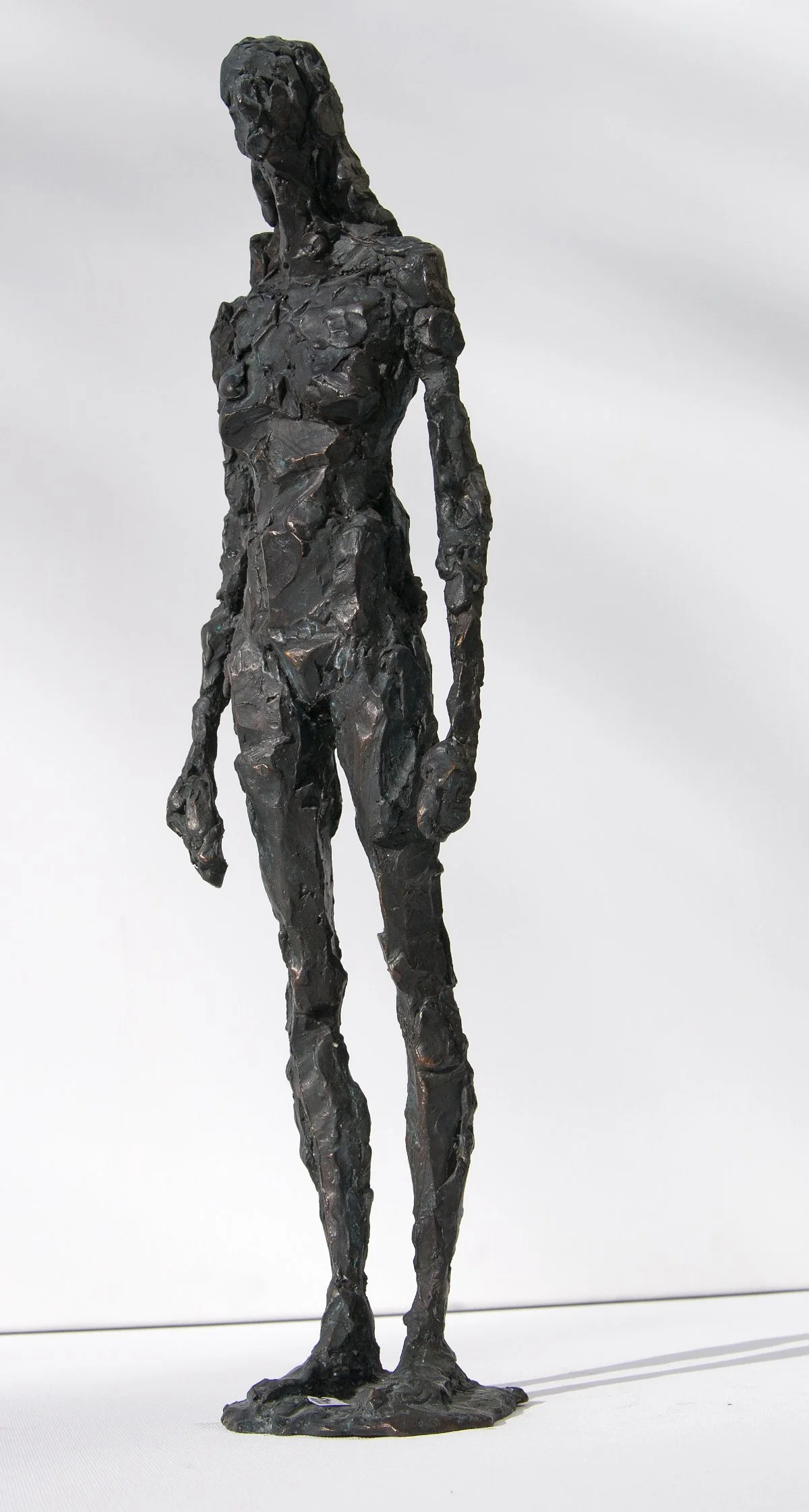 Skulptur einer hohen, abstrakten menschlichen Figur aus dunklem, strukturiertem Material, die auf einem flachen Sockel vor einem schlichten weißen Hintergrund steht.
