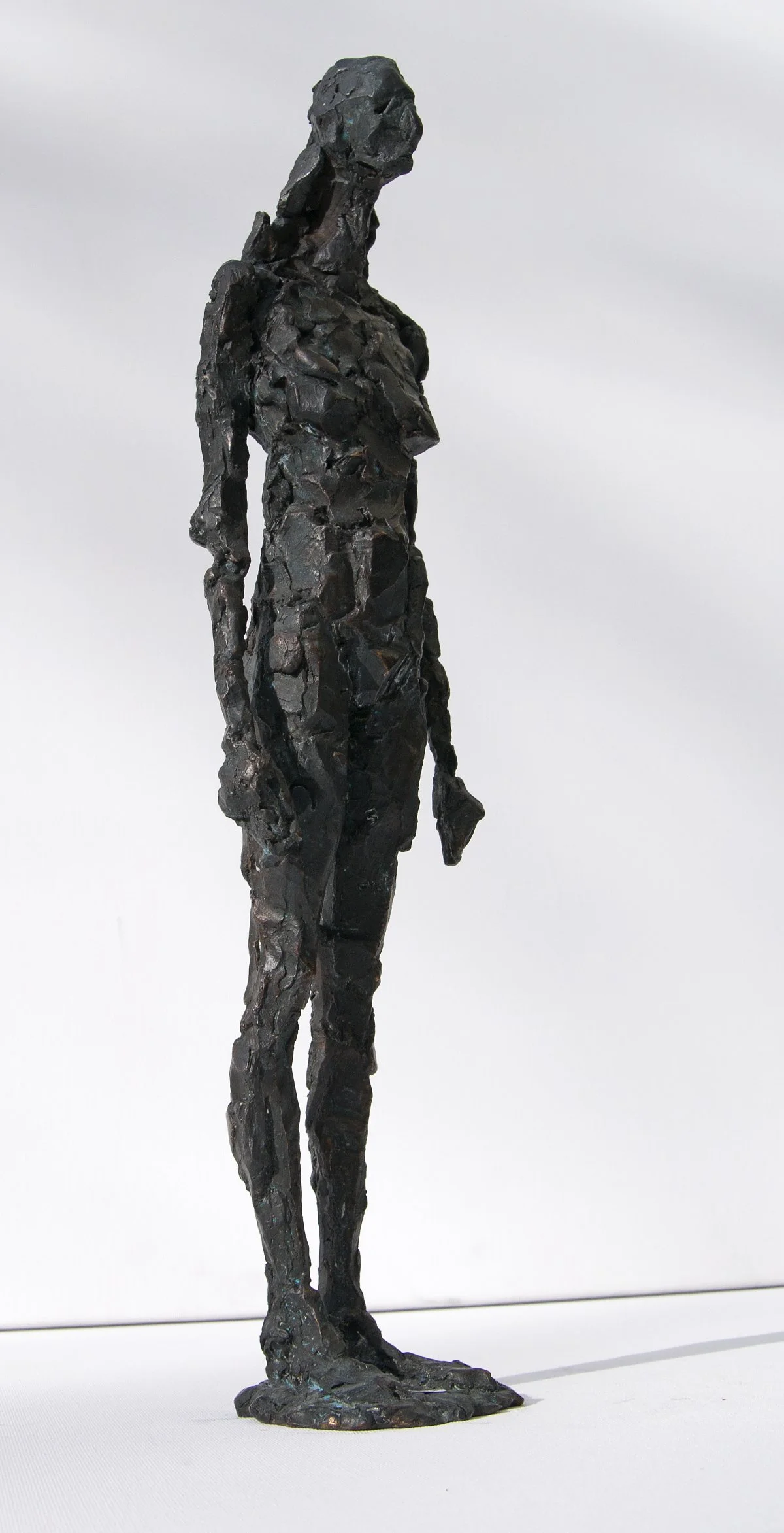 Künstlerische Skulptur einer schlanken weiblichen Figur aus dunklem, strukturiertem Material, die aufrecht vor einem schlichten hellen Hintergrund steht.
