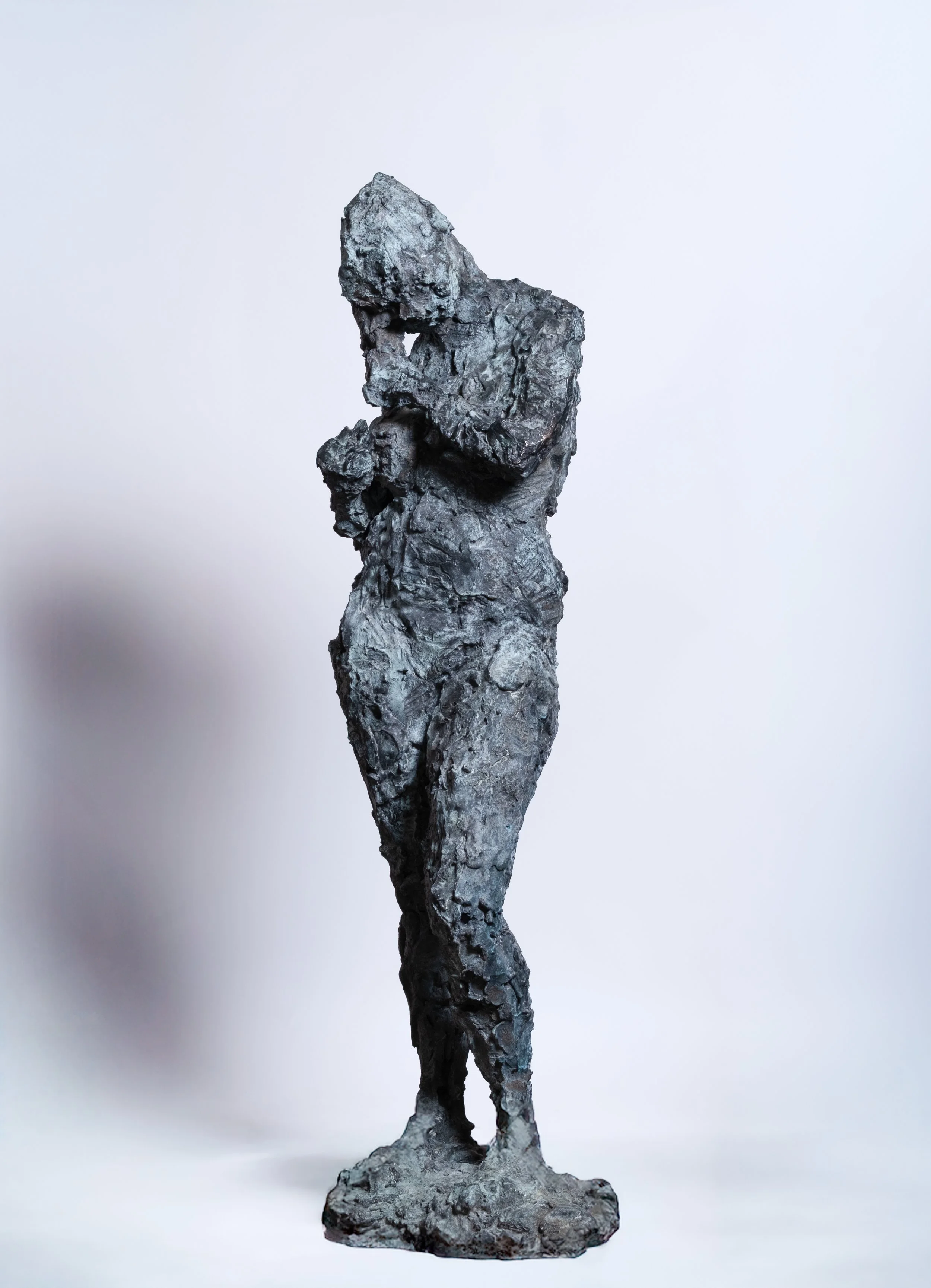 Sculpture of a woman made from rough, textured material, standing with one arm bent and her head slightly bowed, set against a plain light background.
