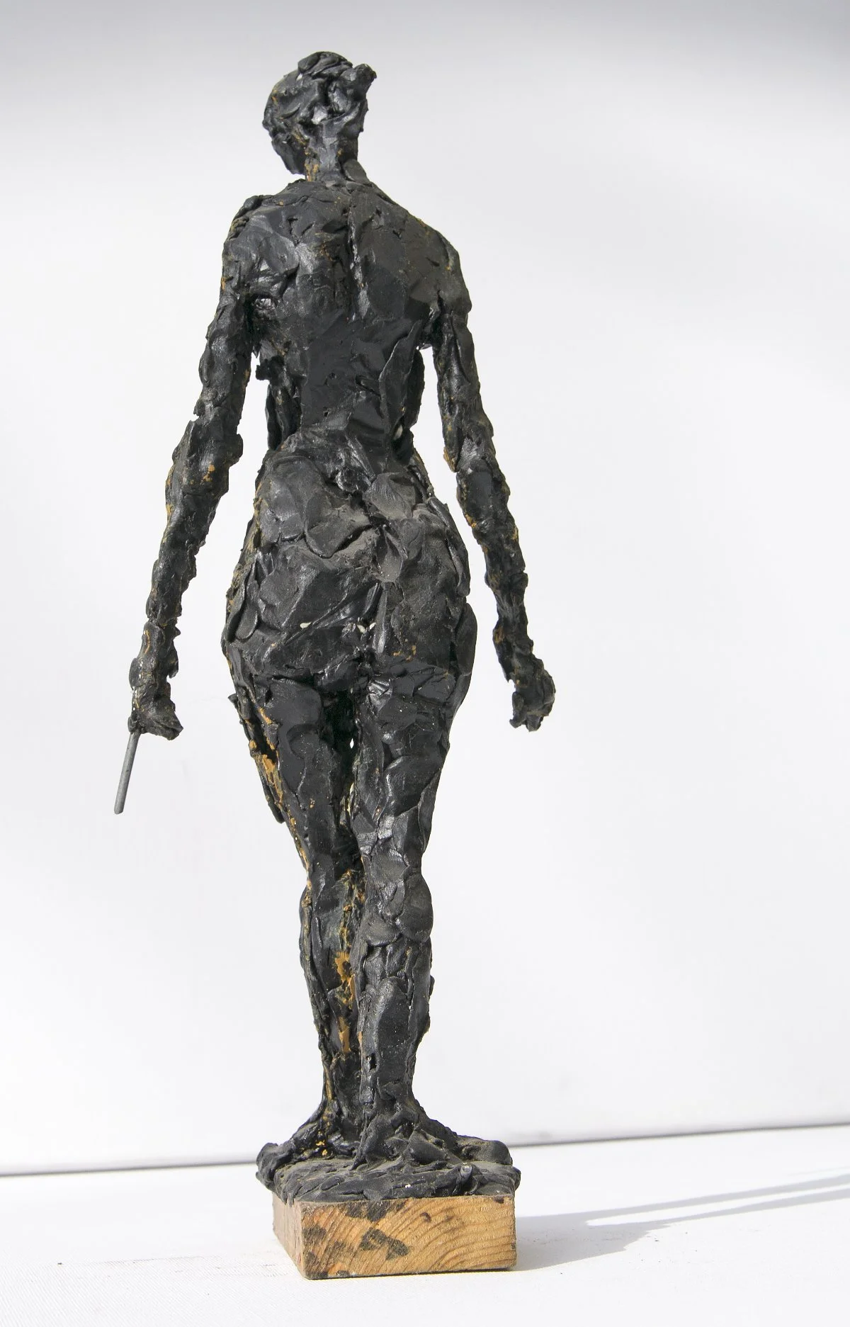 Eine Skulptur einer Frau, die auf einem Holzsockel steht, von hinten gesehen, in ihrer rechten Hand einen Gegenstand hält, mit kurzem Haar und einem detailliert strukturierten Körper.
