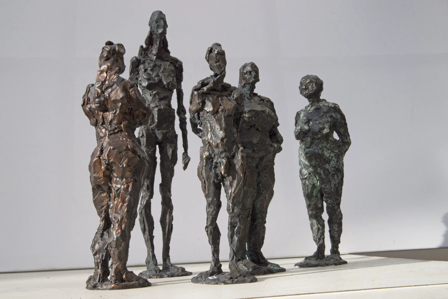 Eine Skulptur aus fünf abstrahierten menschlichen Figuren, jede mit eigener Körperhaltung und -form, gefertigt aus strukturiertem, dunklem Metall, präsentiert auf einer weißen Fläche vor einem schlichten Hintergrund.
