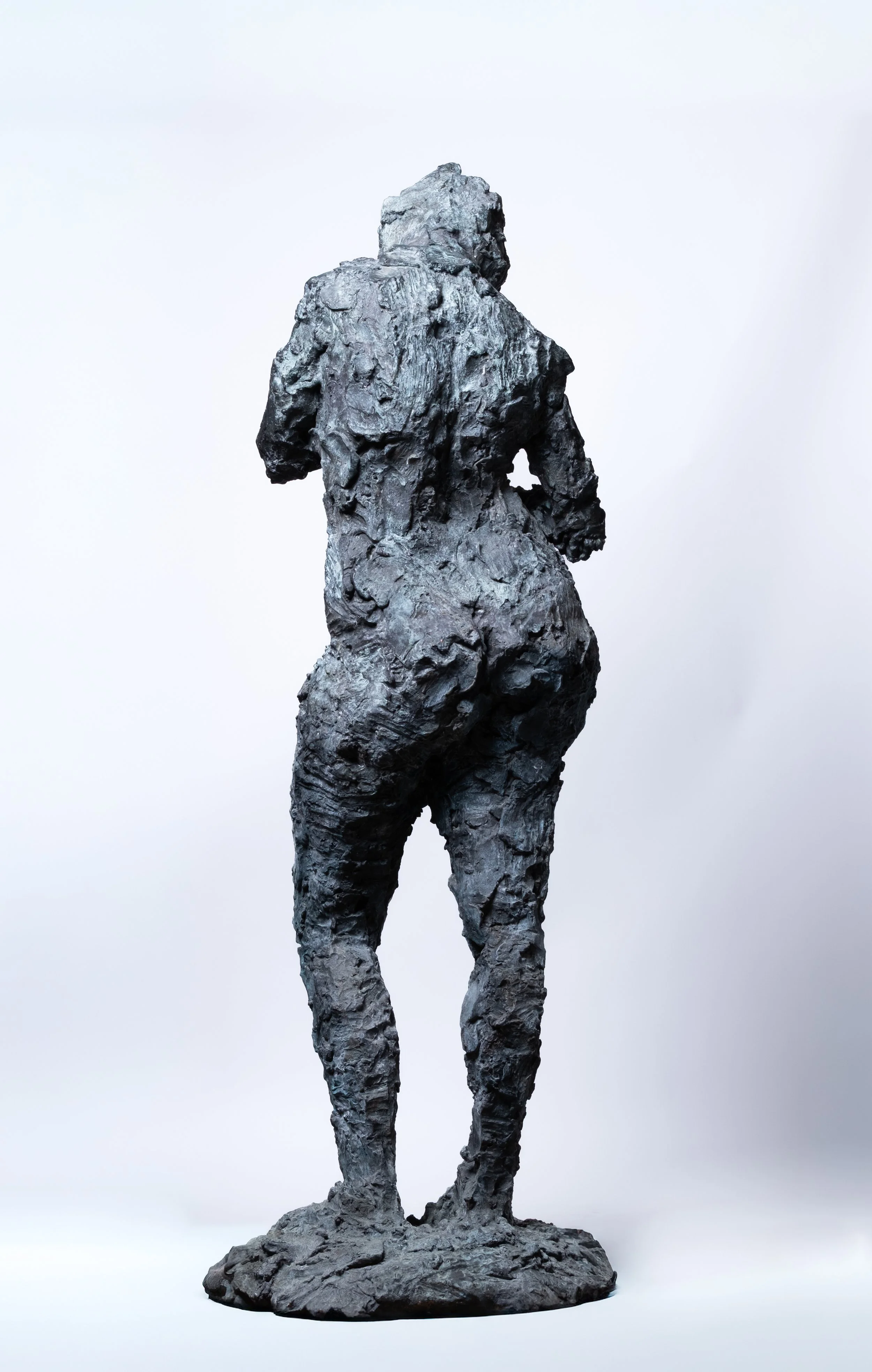 Skulptur einer Frau von hinten, aus einem rauen, strukturierten Material, stehend auf einem Sockel.