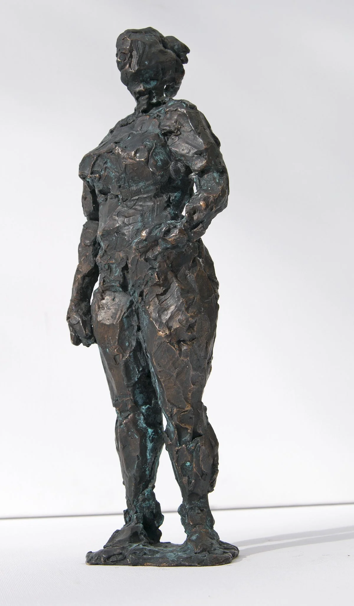 Eine Skulptur einer Frau, die mit in die Hüften gestemmten Händen dasteht, strukturiert und abstrakt im Stil, vor einem schlichten weißen Hintergrund.
