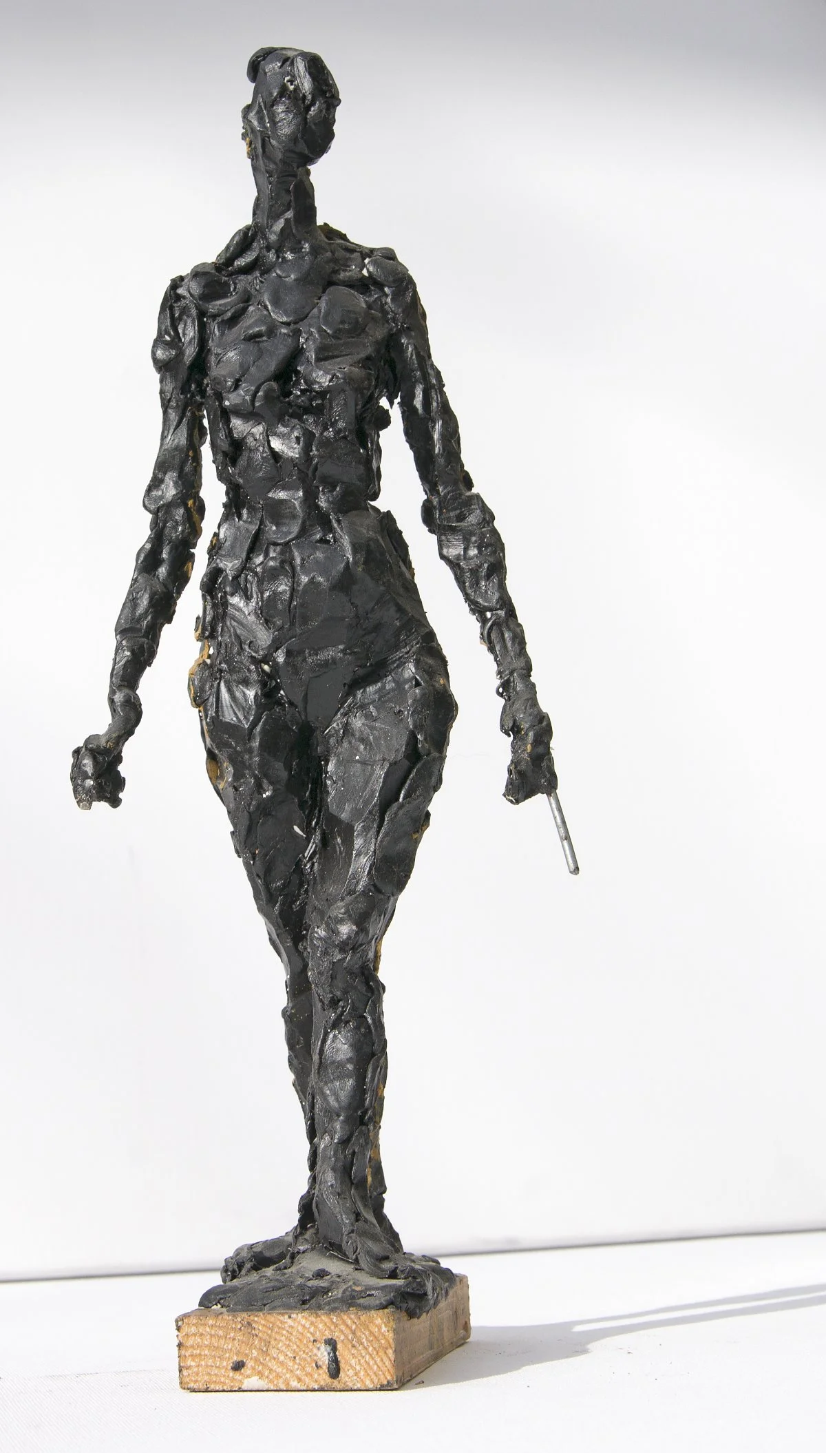 Eine Skulptur einer schlanken Frau aus dunklem, rauem Material, die auf einem kleinen Holzsockel vor einem schlichten weißen Hintergrund steht.
