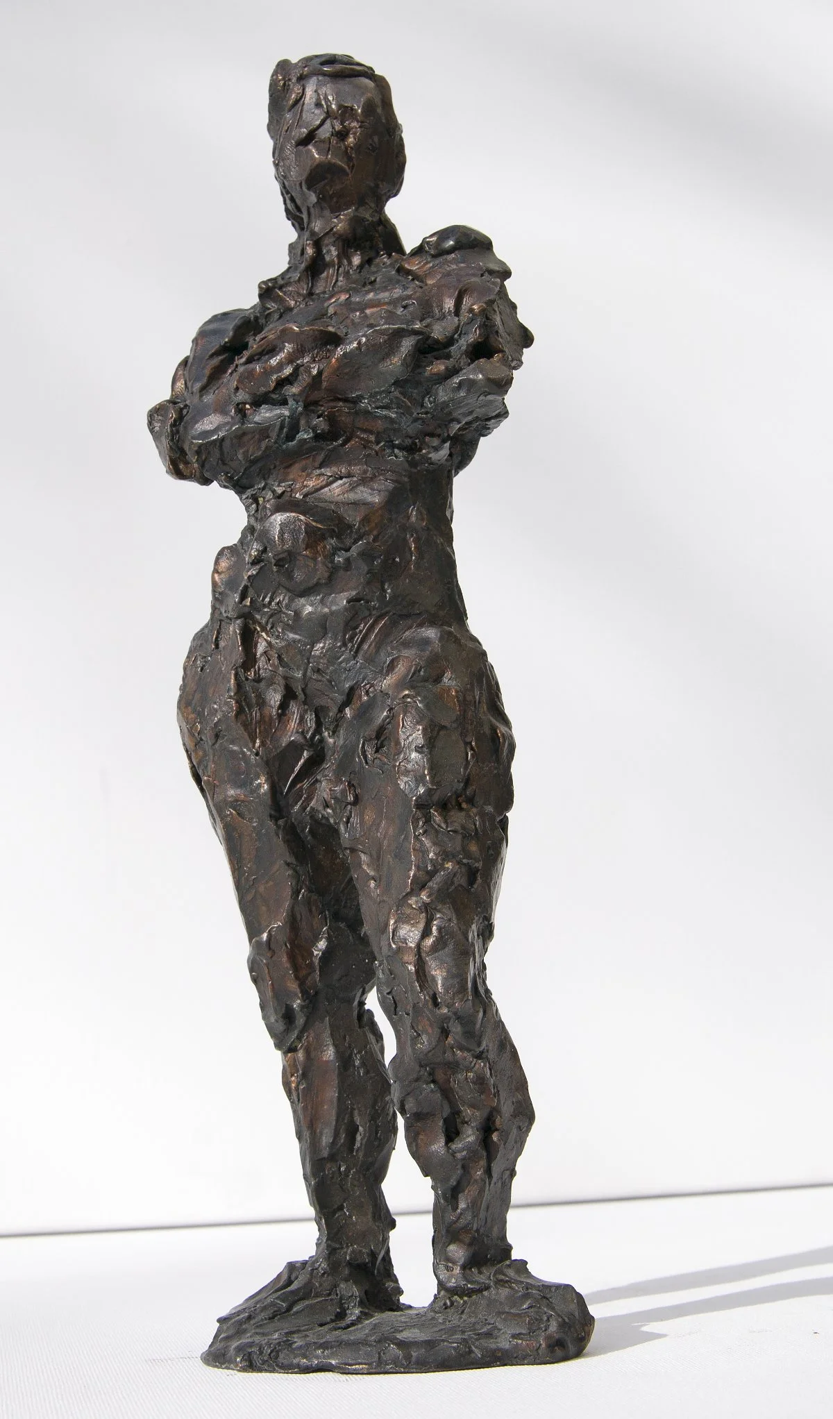 Bronzeskulptur einer Frau mit verschränkten Armen, mit rauer, strukturierter Oberfläche.
