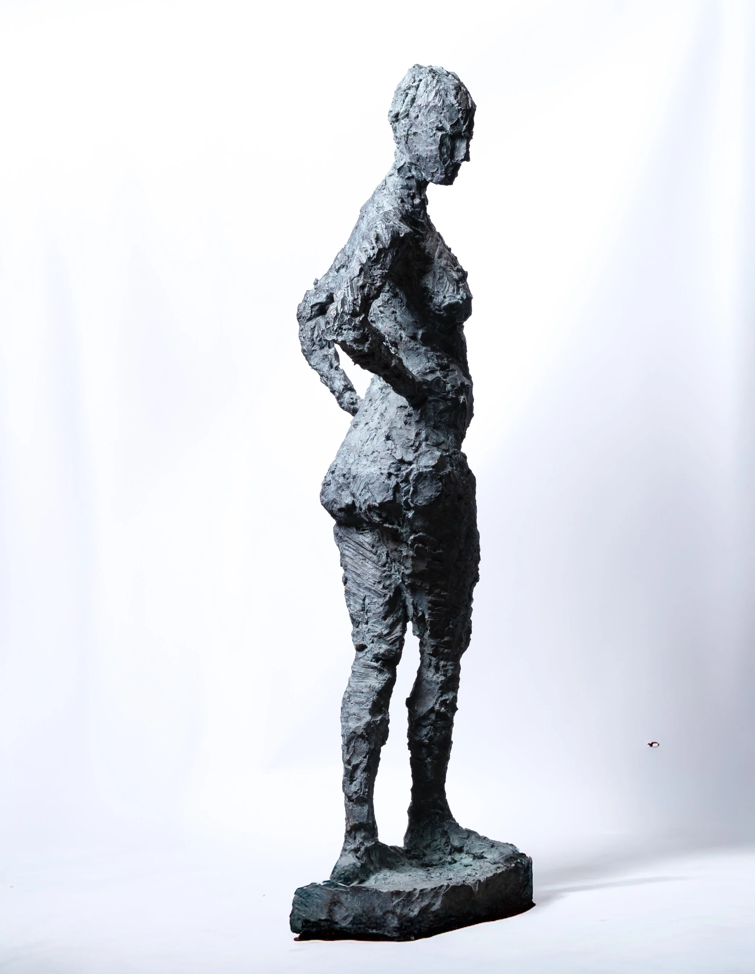 Eine Skulptur einer Frau aus strukturiertem, dunklem Material, die im Profil auf einem rechteckigen Sockel vor einem schlichten weißen Hintergrund steht.