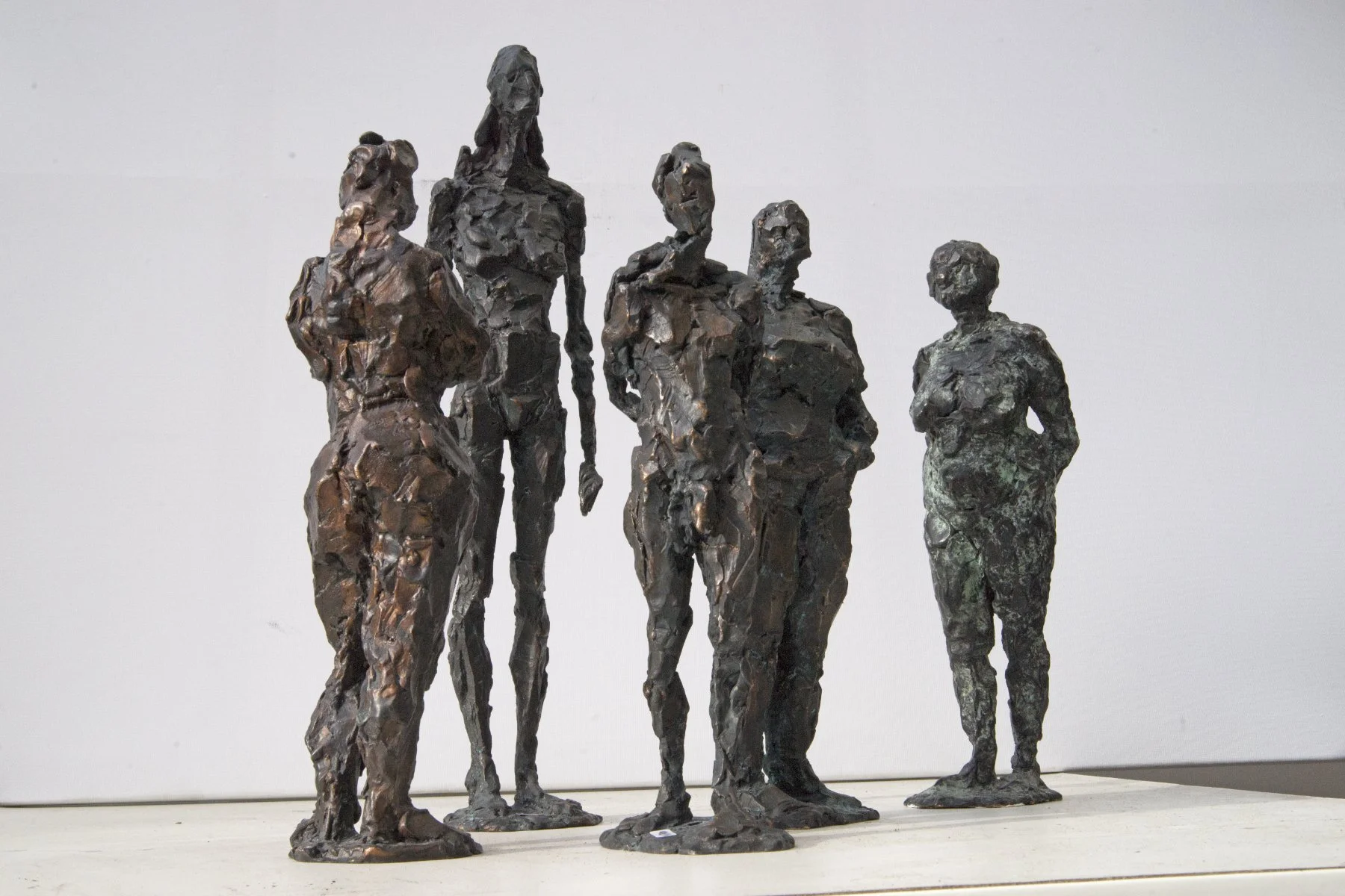 Fünf abstrakte Bronzeskulpturen von Frauen und einem Mann, die auf einer weißen Fläche vor einem schlichten hellen Hintergrund stehen.
