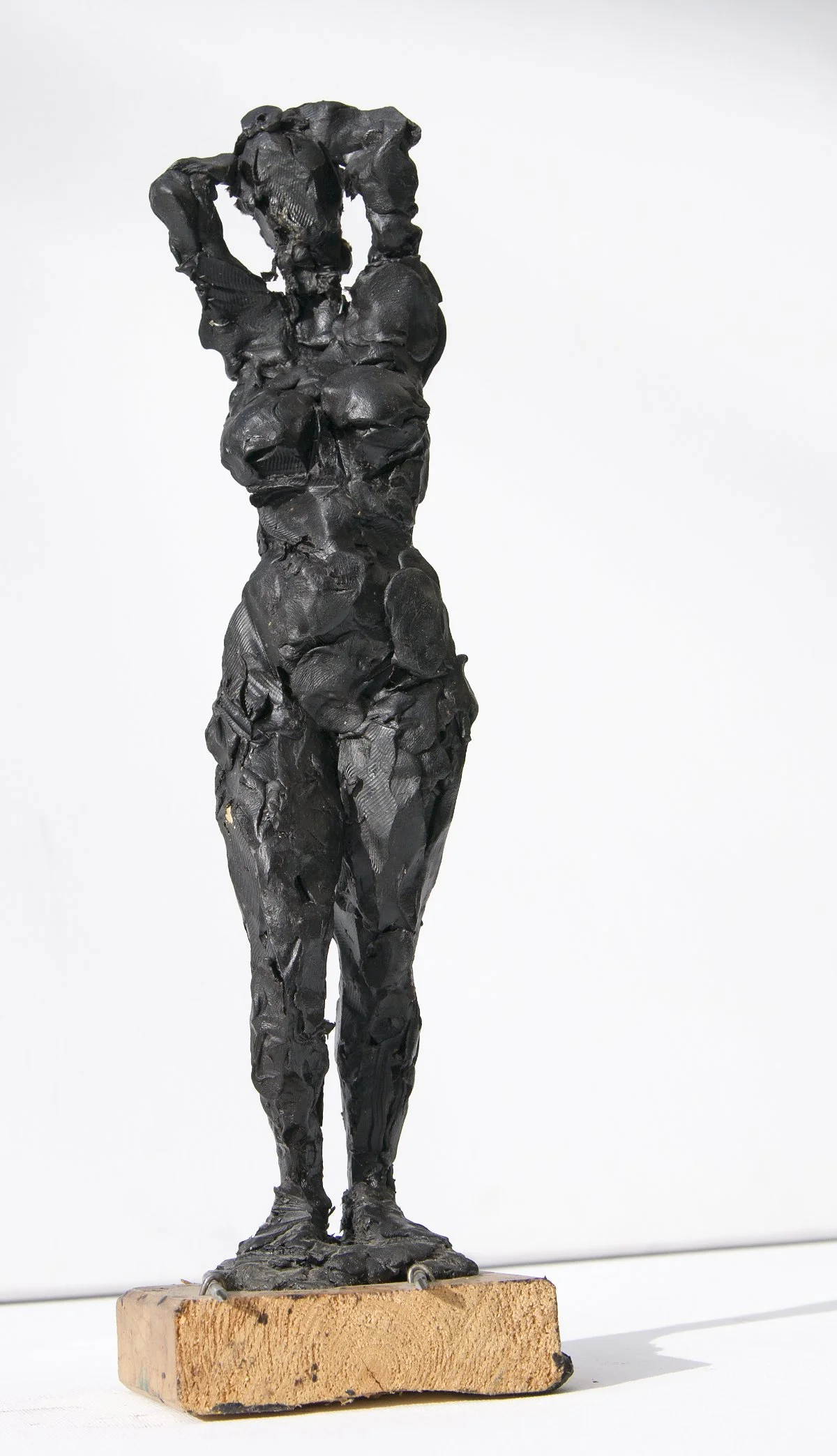 Eine dunkel gefärbte, strukturierte Skulptur einer Frau, die auf einem Holzsockel vor weißem Hintergrund steht und ihre Hand auf dem Kopf ruhen lässt.
