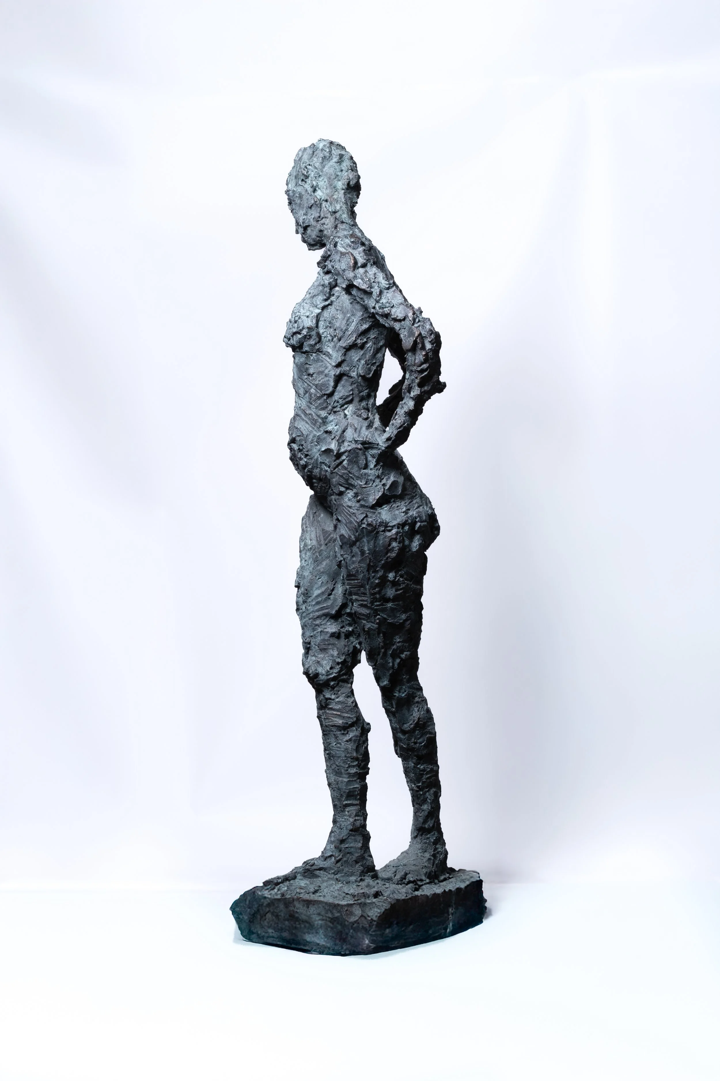 Sculpture of a standing woman made of textured dark material on a small base, posed with one hand on her hip, against a plain white background.