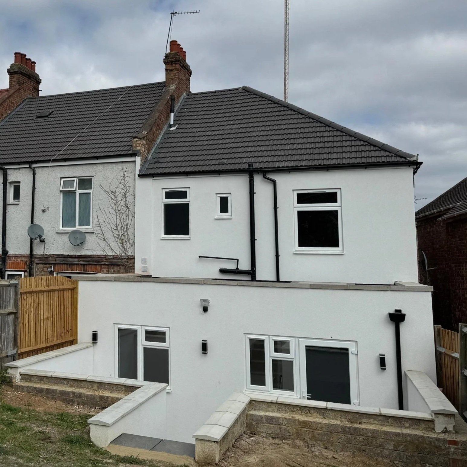 Two-story house with white exterior walls, black roof tiles, and multiple windows under a cloudy sky. The house has a new extension at the back, with a lower level featuring large sliding glass doors and a staircase leading down to a garden area. A w