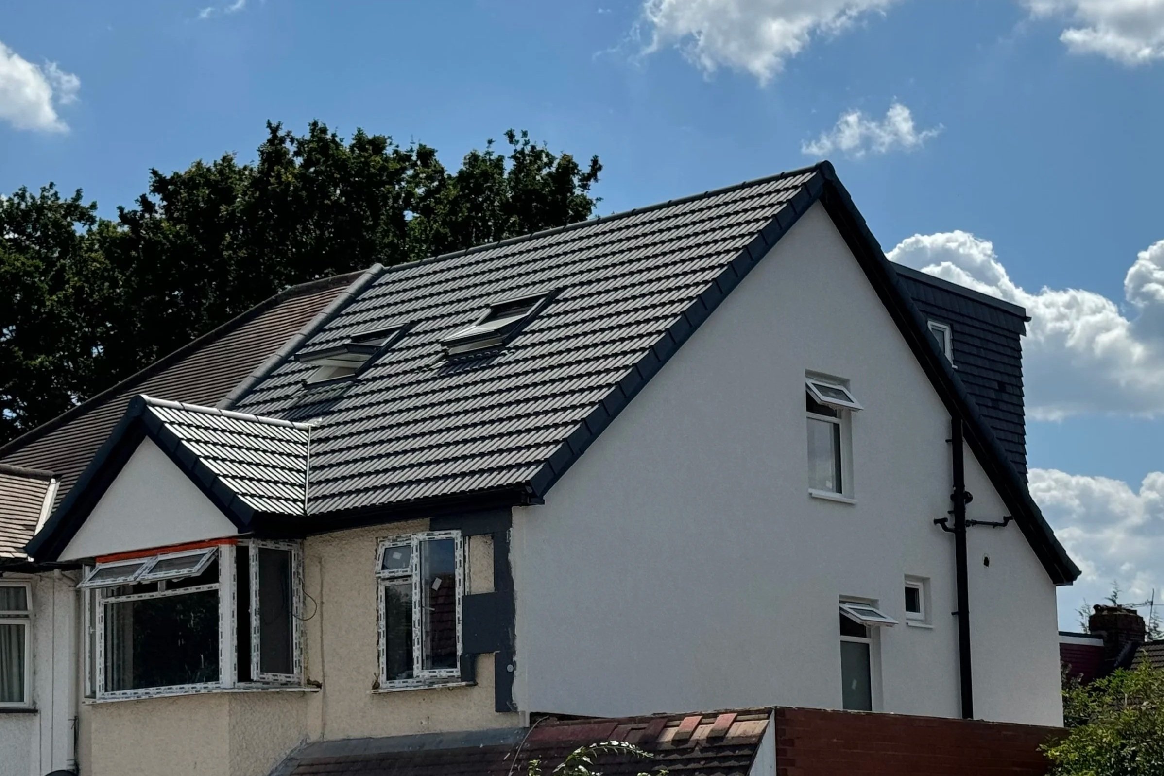 A white two-story house with a dark gray sloped roof and multiple windows, some of which are open. The background shows a partly cloudy sky and green trees.