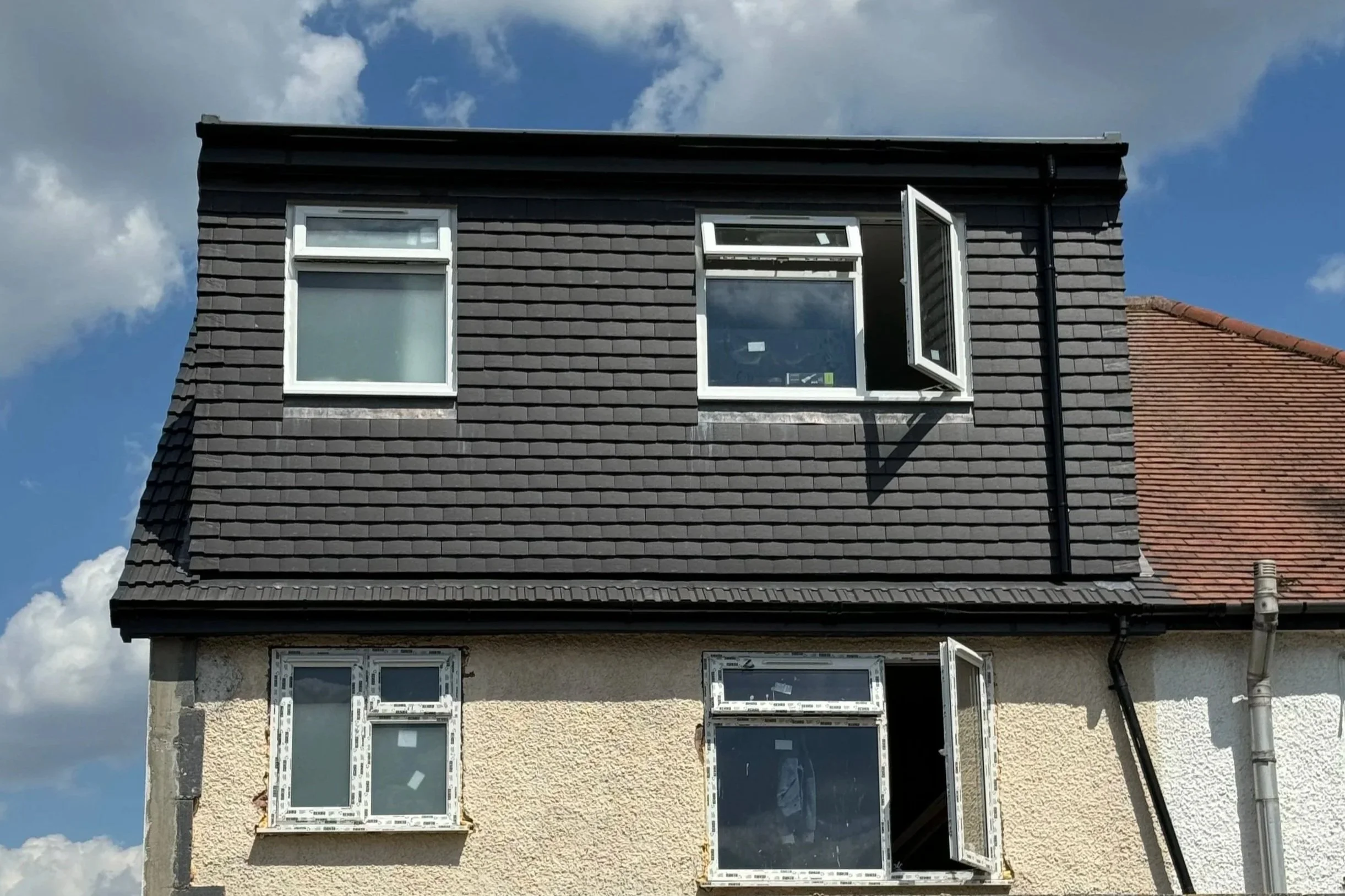 Partially constructed house with beige stucco on the lower part and black shingles on the upper part. The house has two openings where windows are being installed, with one window open. The roof is seen in the background, with a blue sky and some clouds.