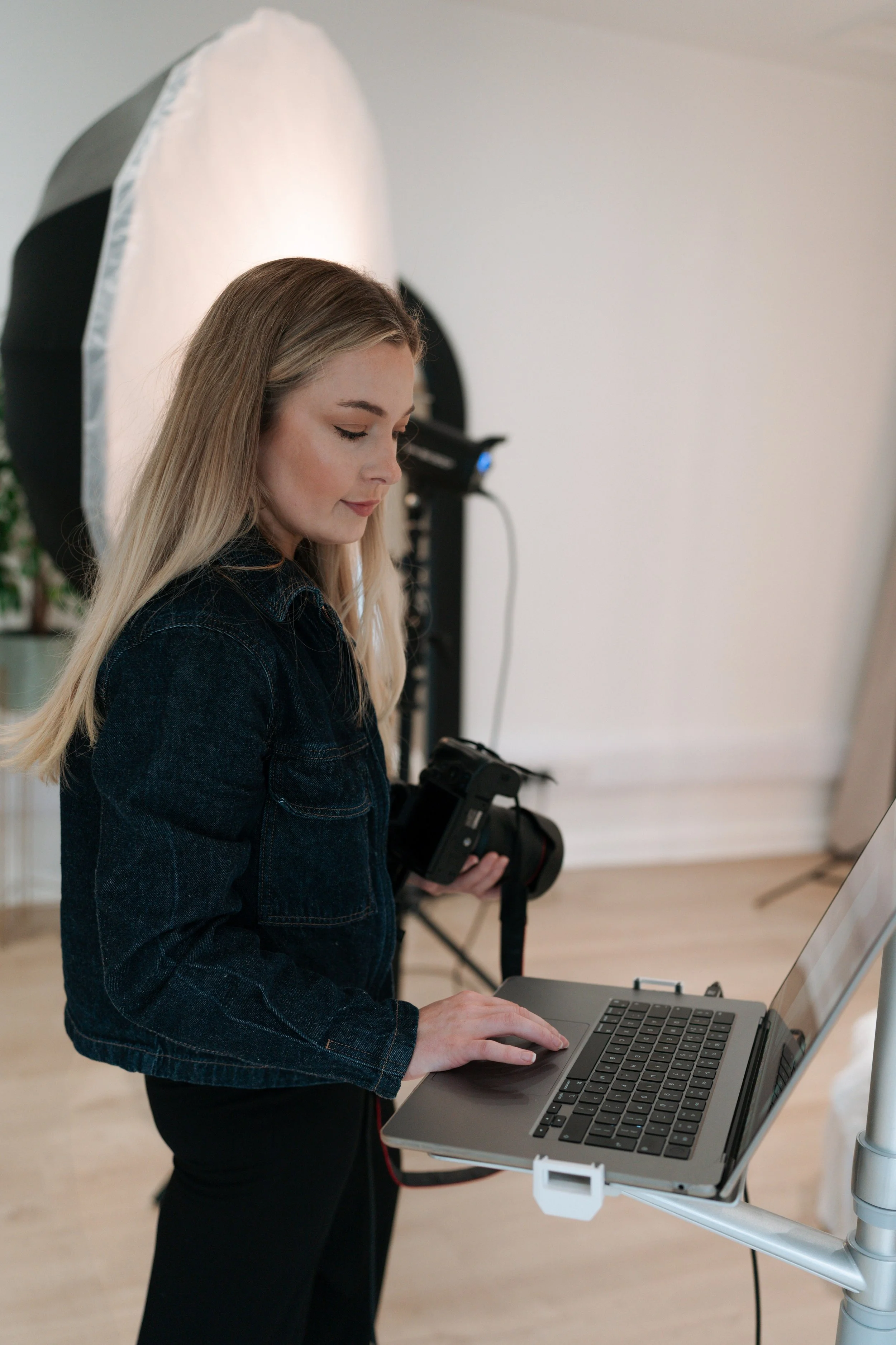 Young woman with blonde hair using a laptop in a photography studio, holding a camera in her right hand.