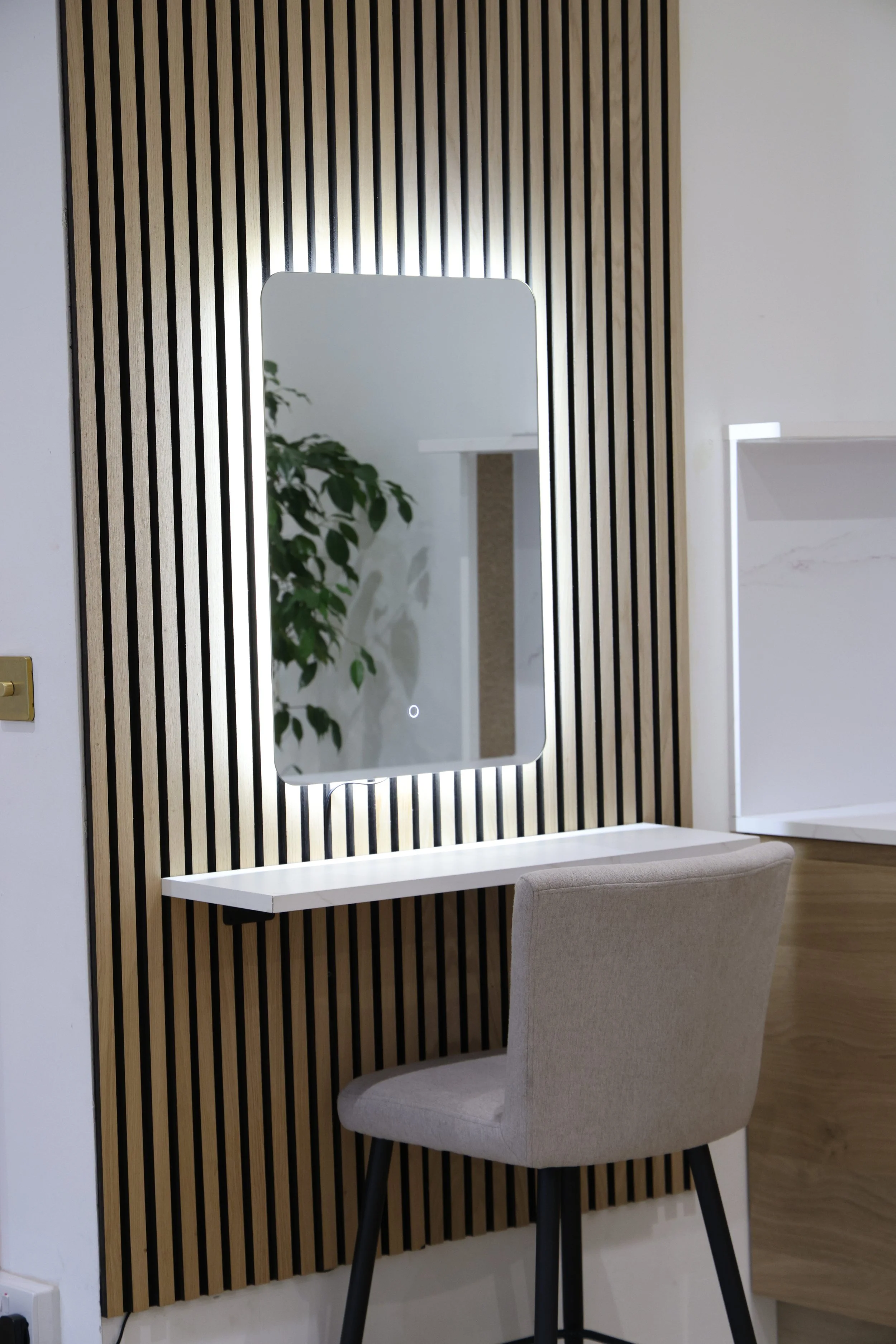 Modern vanity area with a backlit mirror, a white floating desk, a neutral-colored upholstered chair, and a wood-paneled wall.