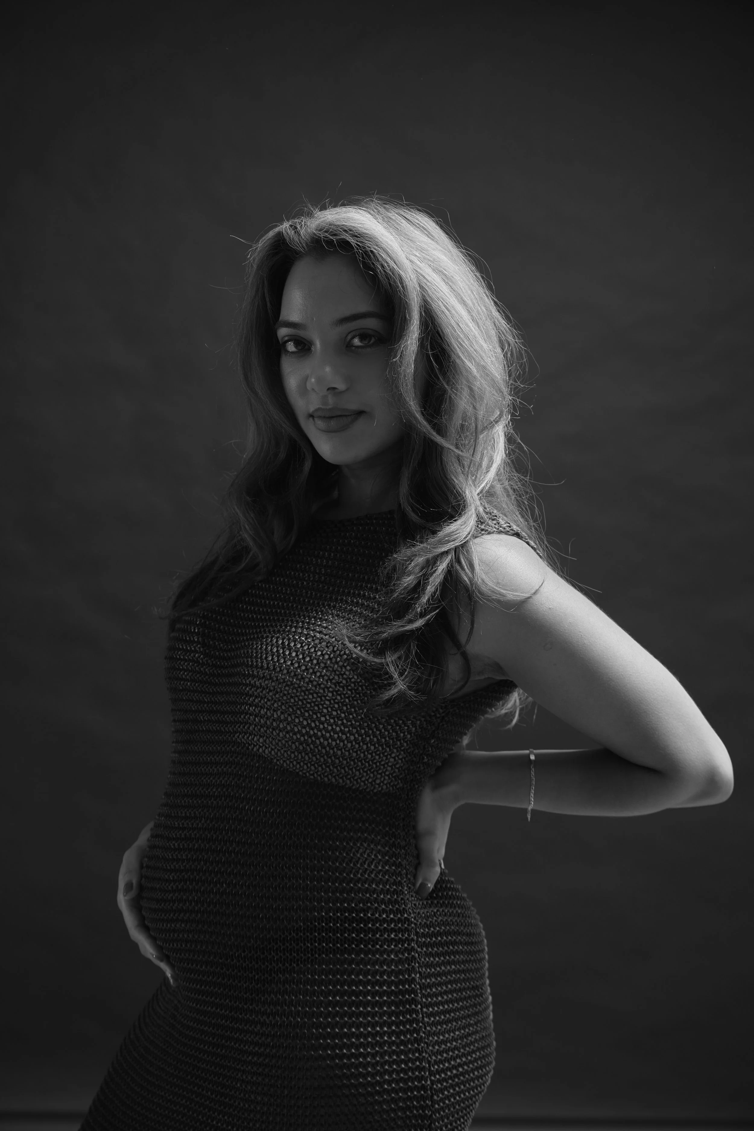Black and white portrait of a woman with long, wavy hair, wearing a textured sleeveless dress, standing with her left hand on her hip and looking at the camera, against a dark plain background.