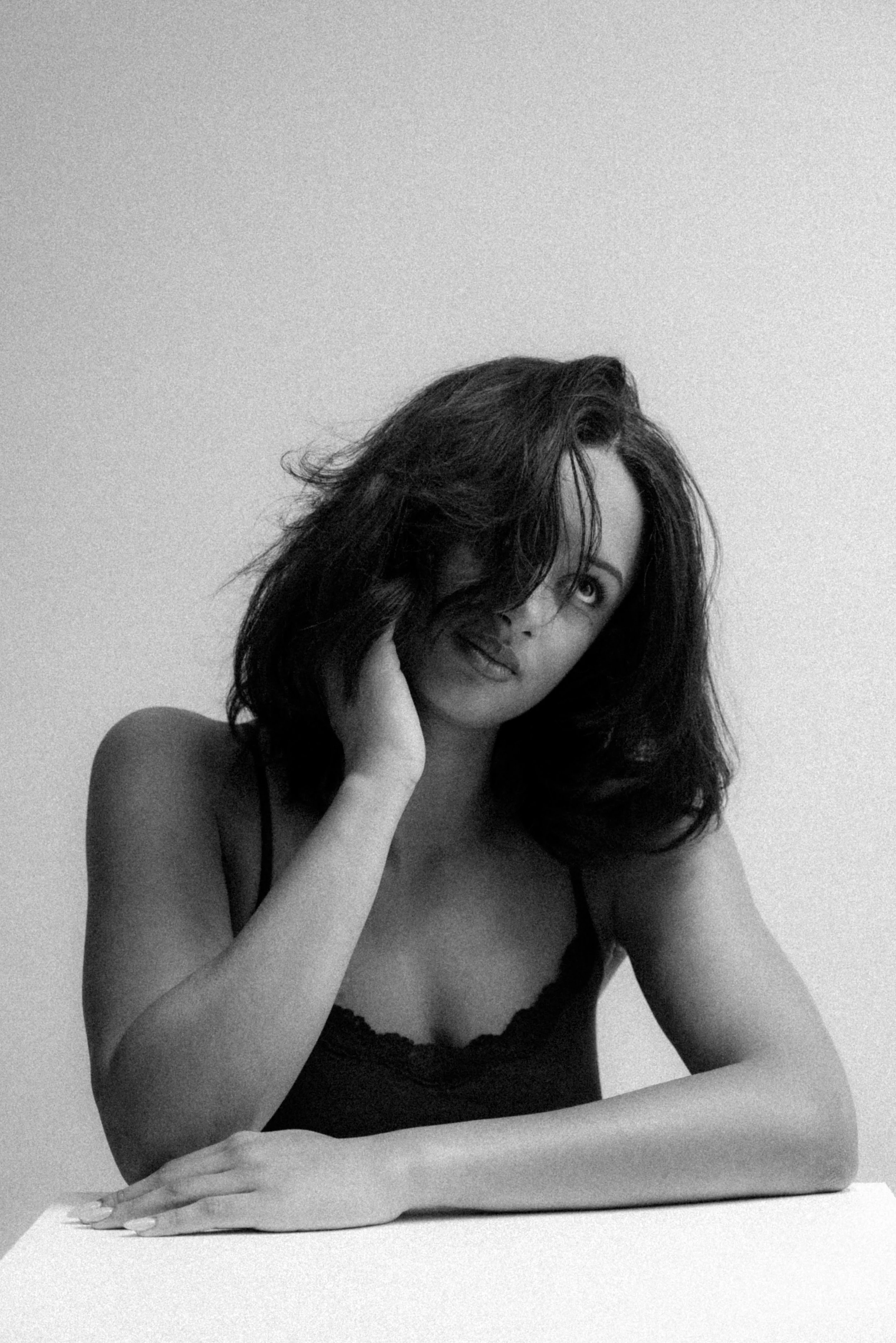 A woman with shoulder-length dark hair, wearing a spaghetti strap top, sitting at a table with her head resting against her hand, looking at the camera with a slight smile.