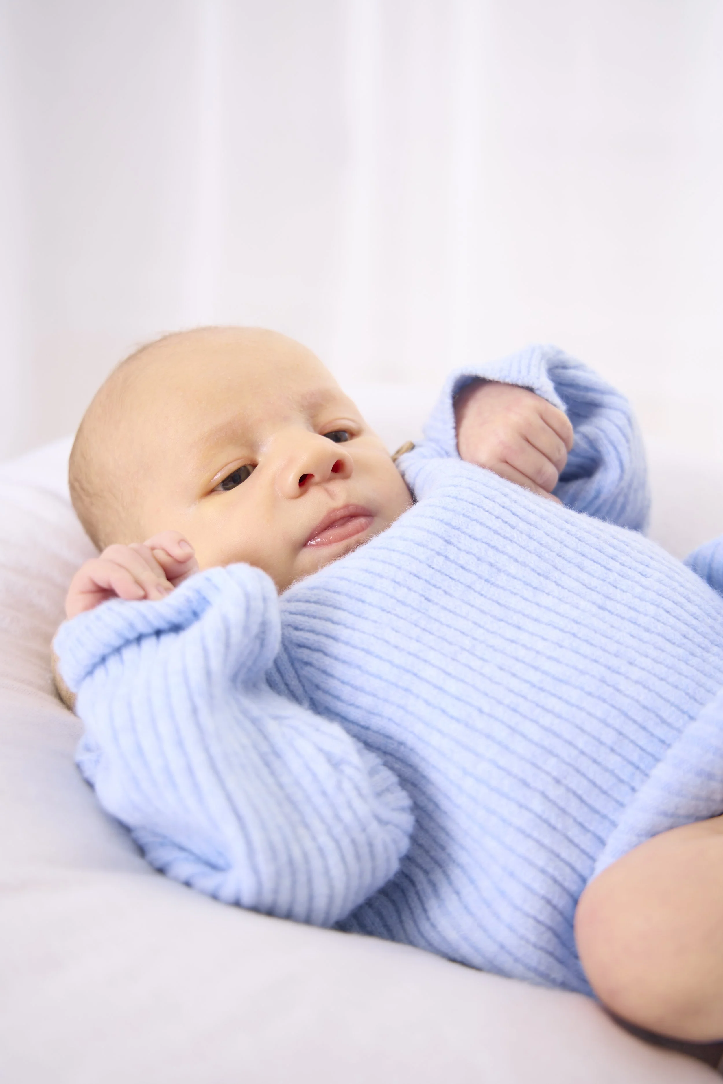 A baby lying on a bed, wearing a light blue knitted sweater, looking relaxed with hands near face.