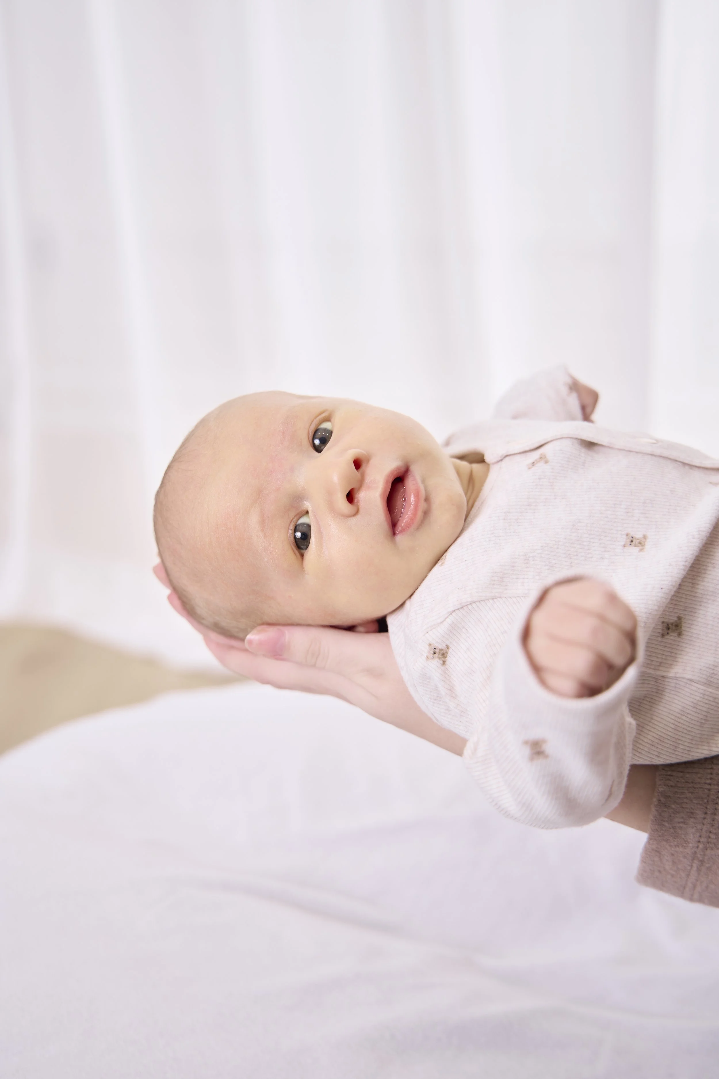 A baby lying on a white surface being gently held by an adult's hand, looking curiously at the camera with a slight smile.
