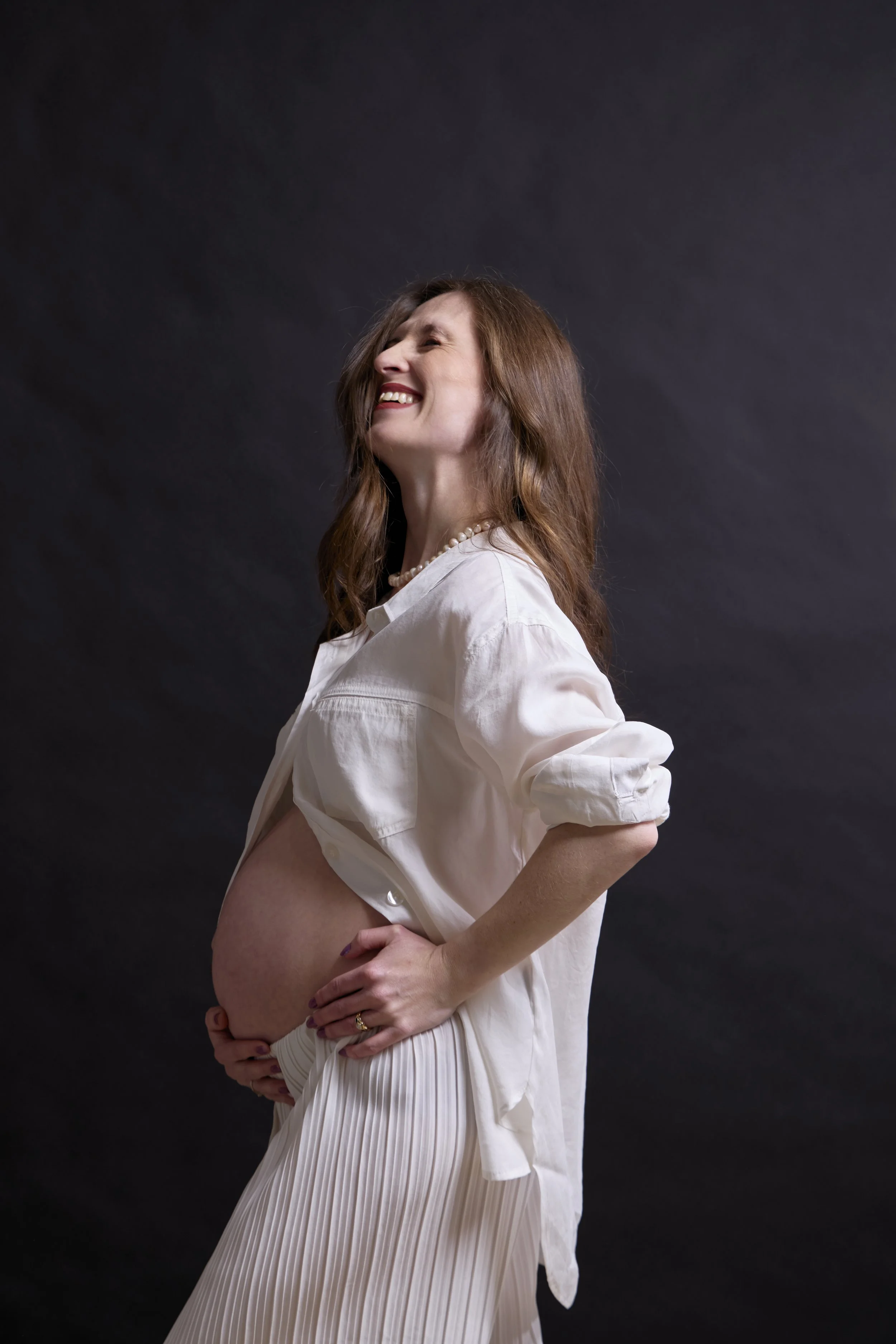 A pregnant woman with long brown hair smiling and looking to her left, wearing a white shirt draped over her shoulders and a pleated white skirt, standing against a dark background.