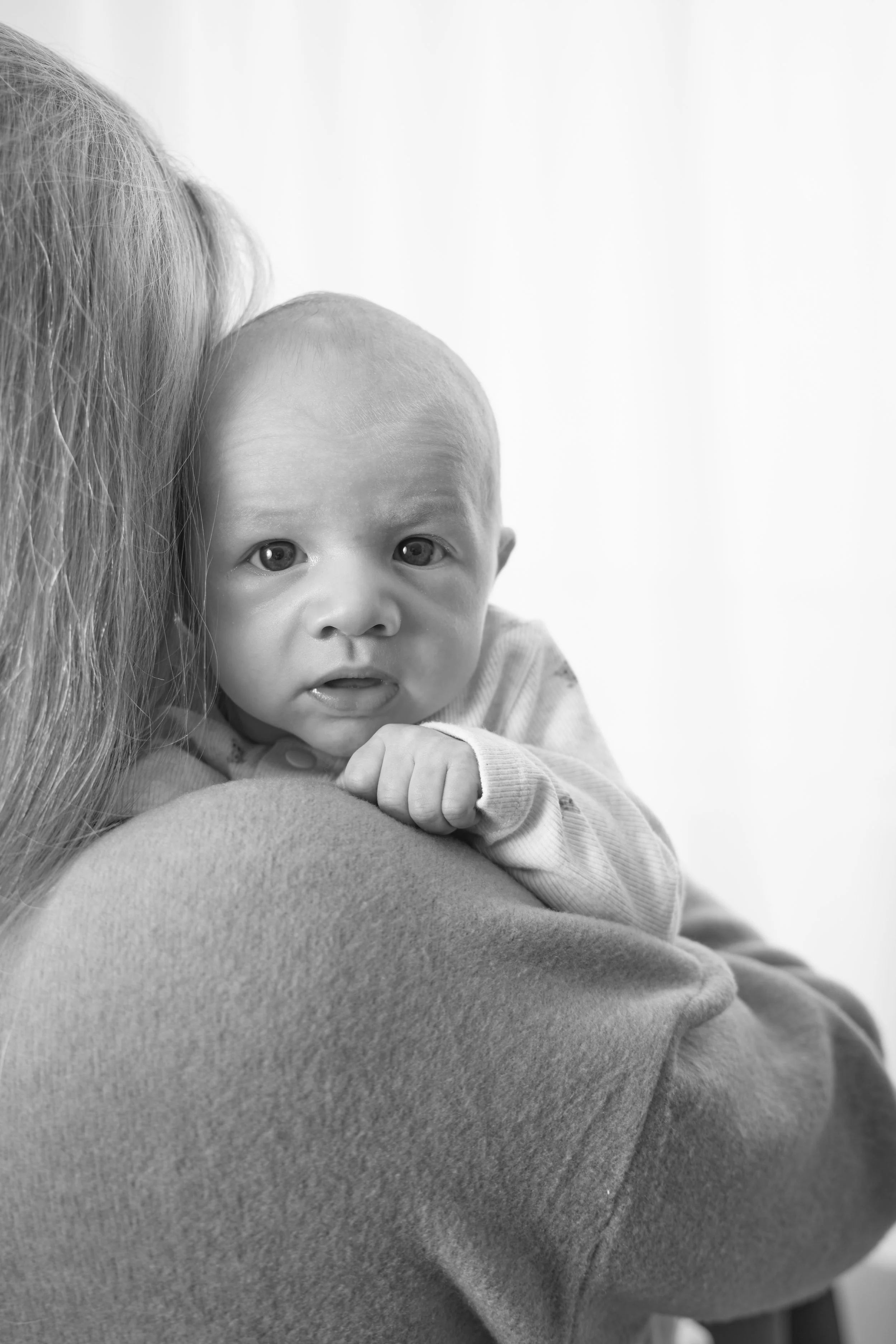 Close-up of a baby with blonde hair and expressive eyes, resting on an adult's shoulder, holding onto their arm, in black and white.