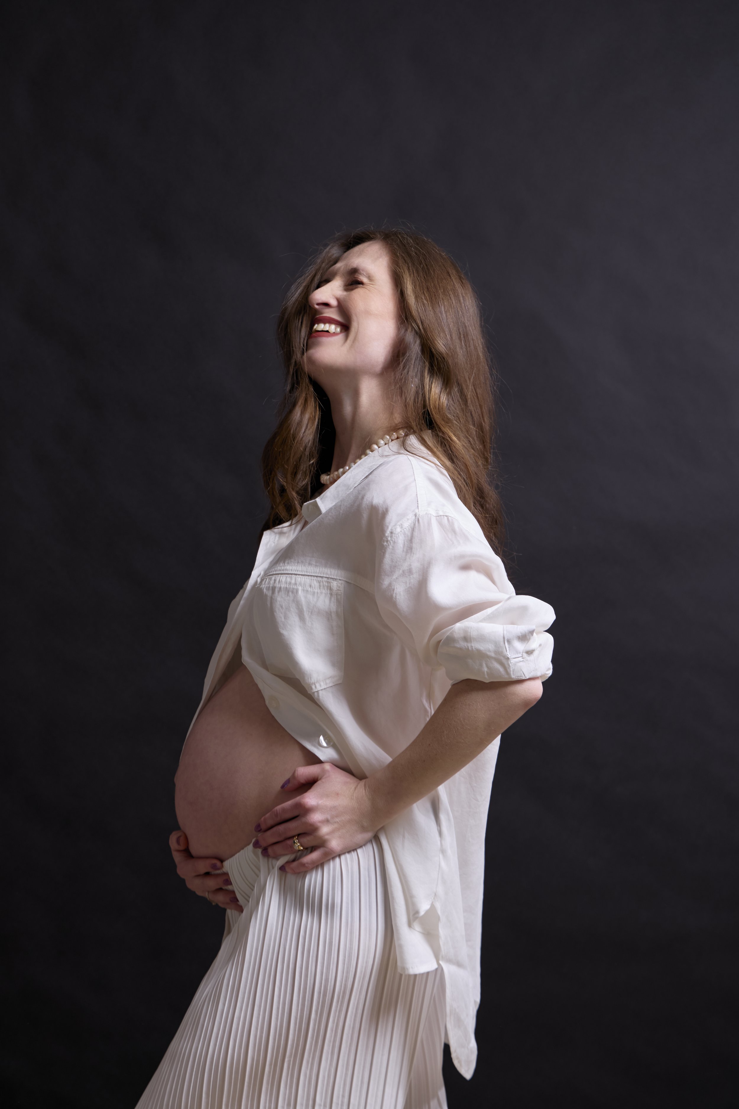 A pregnant woman with long brown hair smiling and standing sideways with her eyes closed, wearing a white shirt and a pleated skirt, against a dark background.