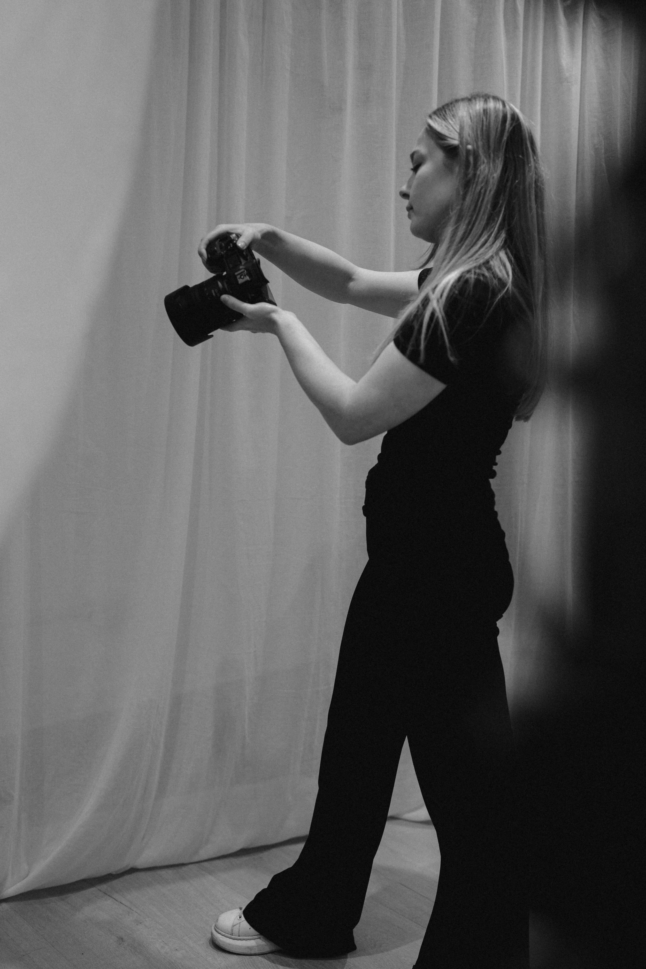 A woman in a black outfit holding a camera, standing next to a curtain.