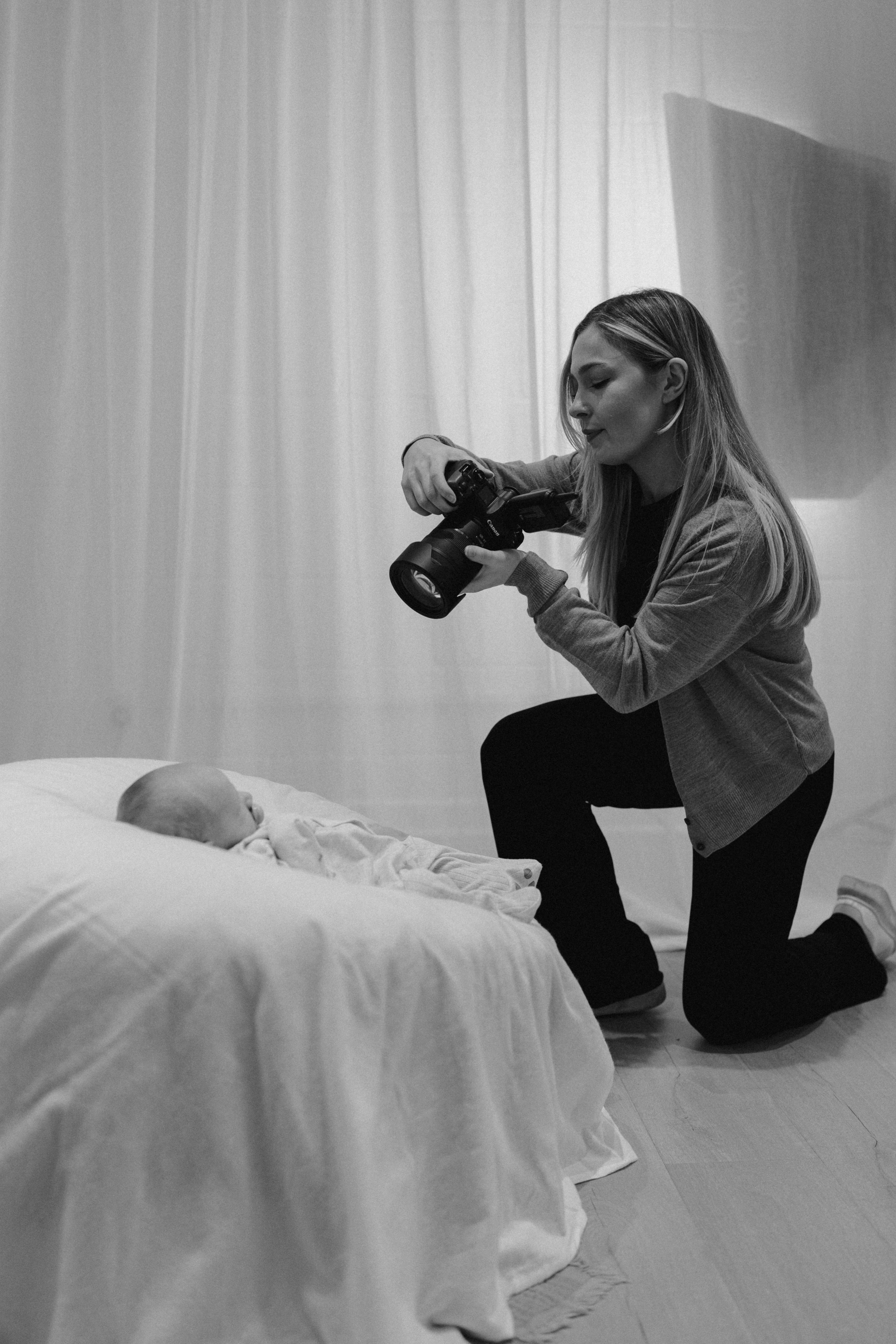 A woman kneels on one knee, holding a camera, taking a photograph of a newborn baby lying on a bed.