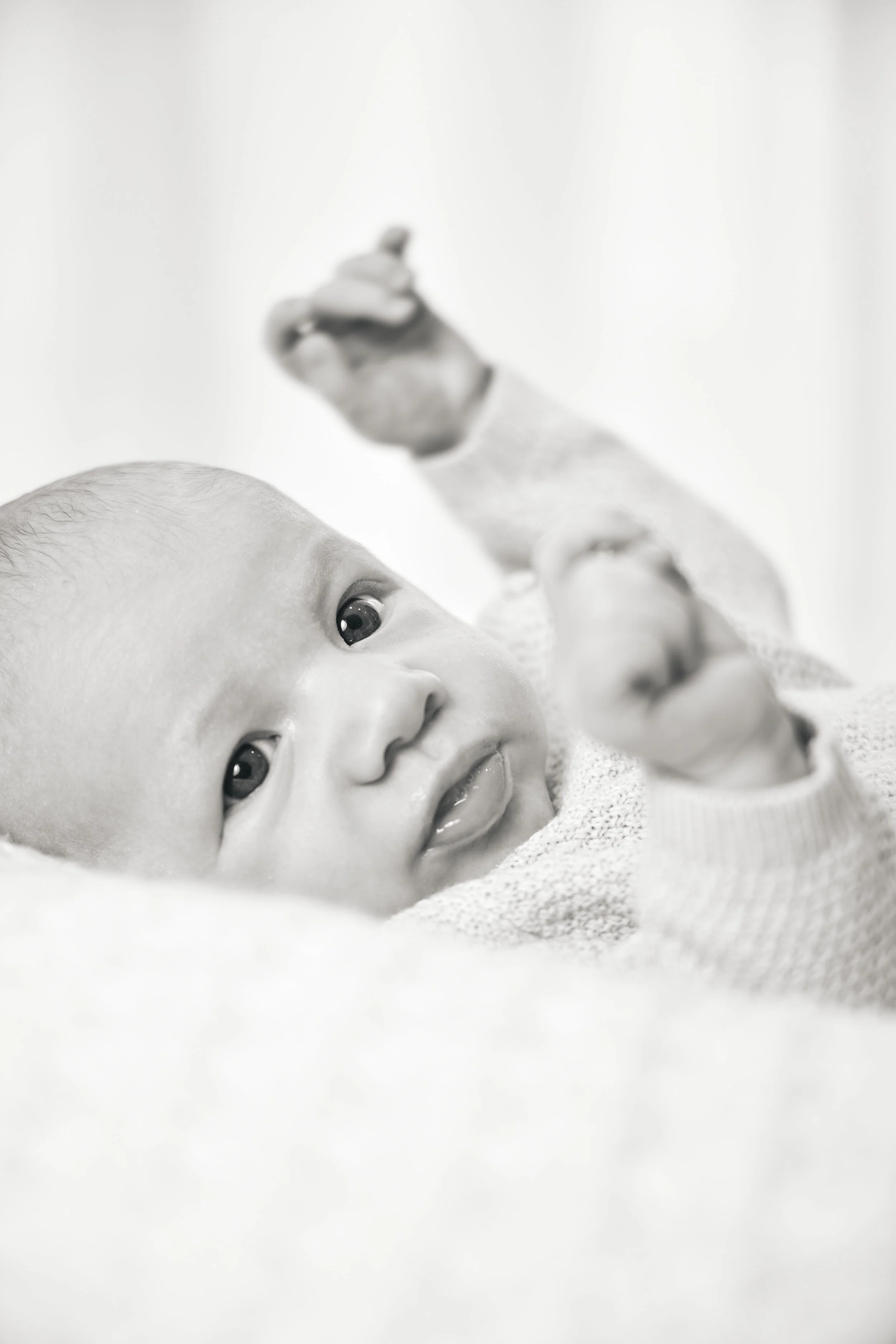 Close-up of a baby lying on a soft surface, looking at the camera with a curious expression, reaching upward with one hand, in black and white.