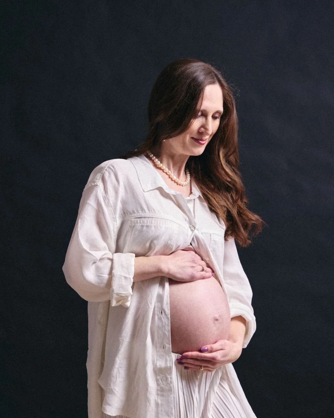 A pregnant woman with long brown hair, wearing a white blouse and pearl necklace, gently holding her belly and looking down affectionately, against a dark background.