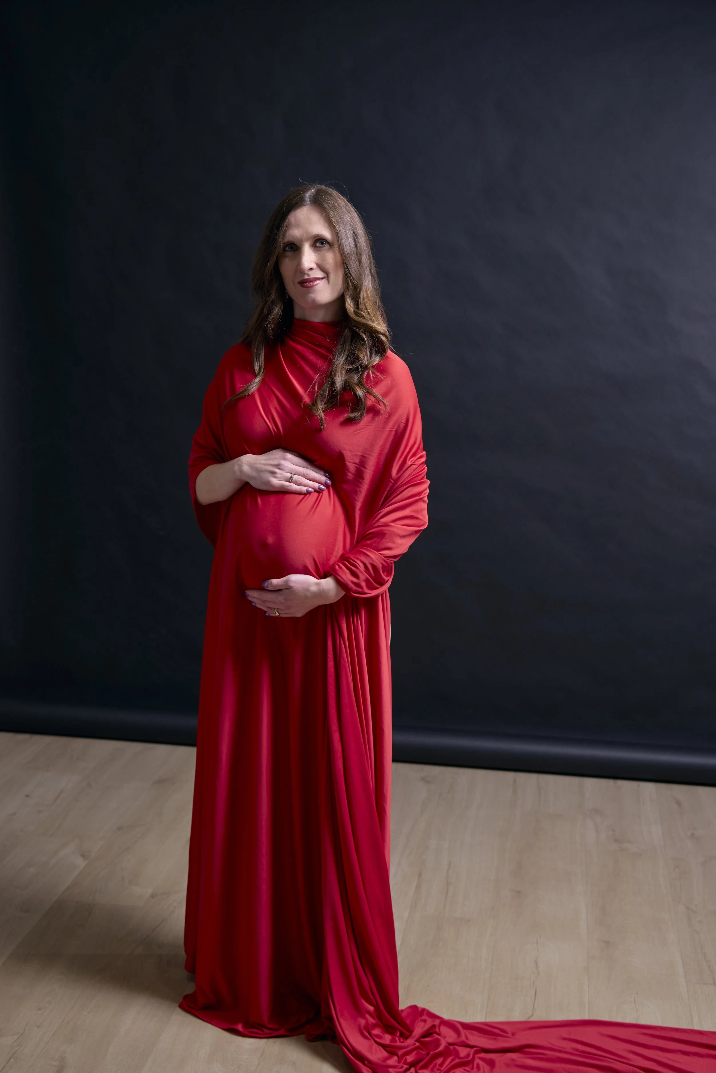 A pregnant woman in a red dress standing on a wooden floor against a dark background, holding her baby bump and smiling.