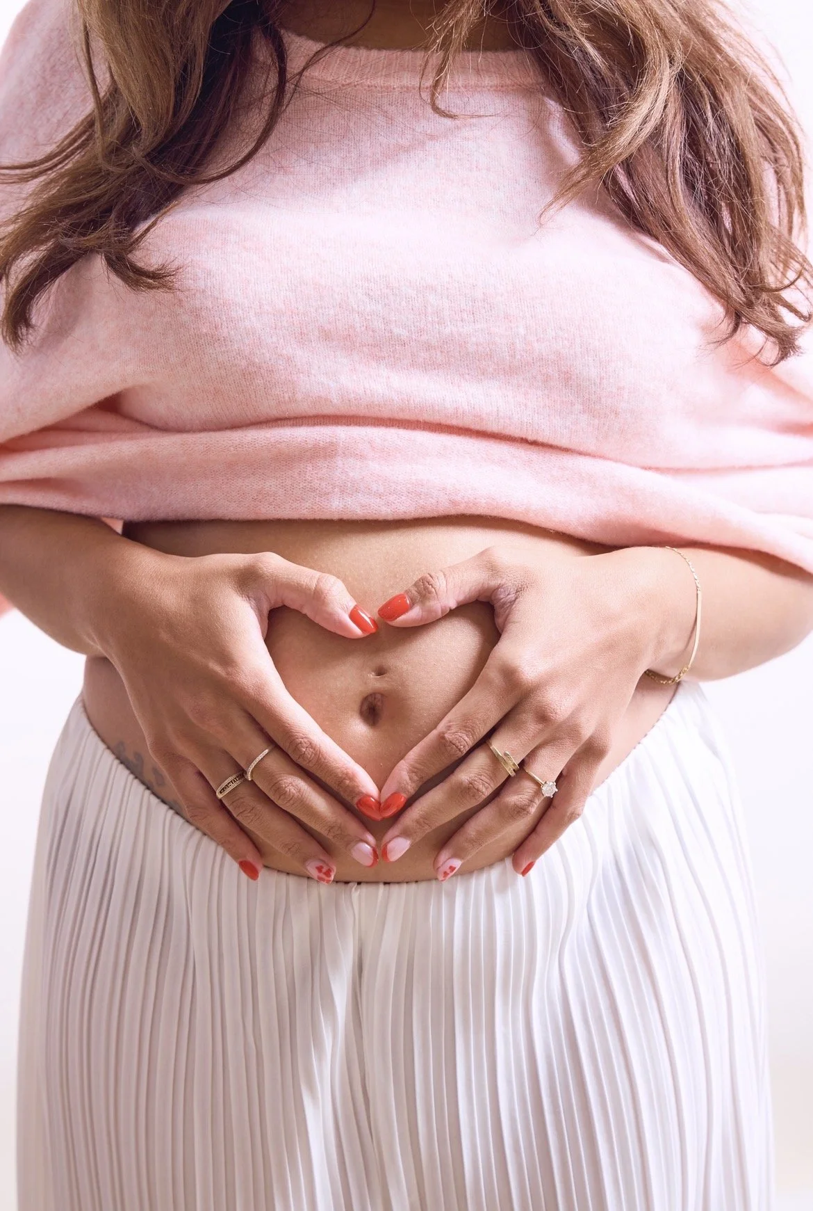 A woman wearing a pink shirt and pleated white skirt forming a heart shape with her hands over her stomach.